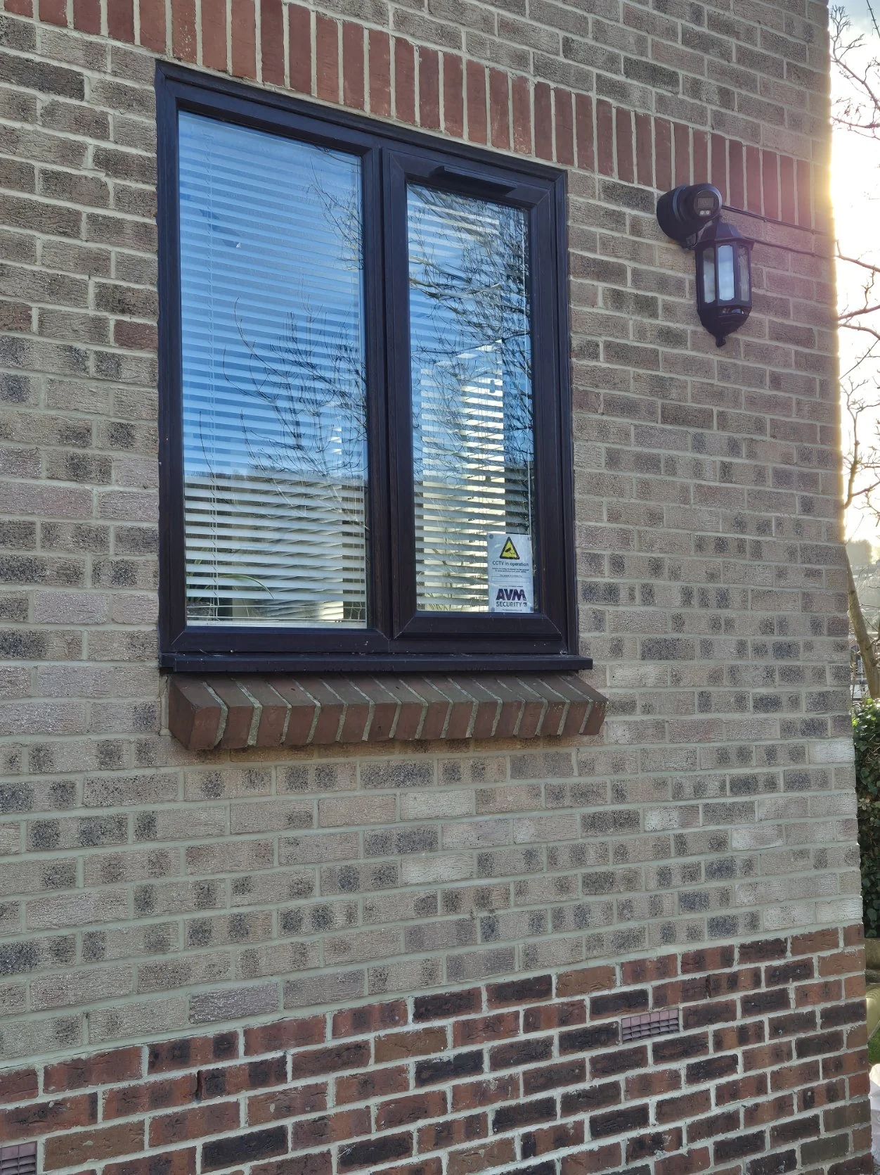 Close-up of a brick house exterior with a double-pane window, black window frame, and tree reflections in the glass. An outdoor wall-mounted light fixture is beside the window, with sunlight coming from the right side.