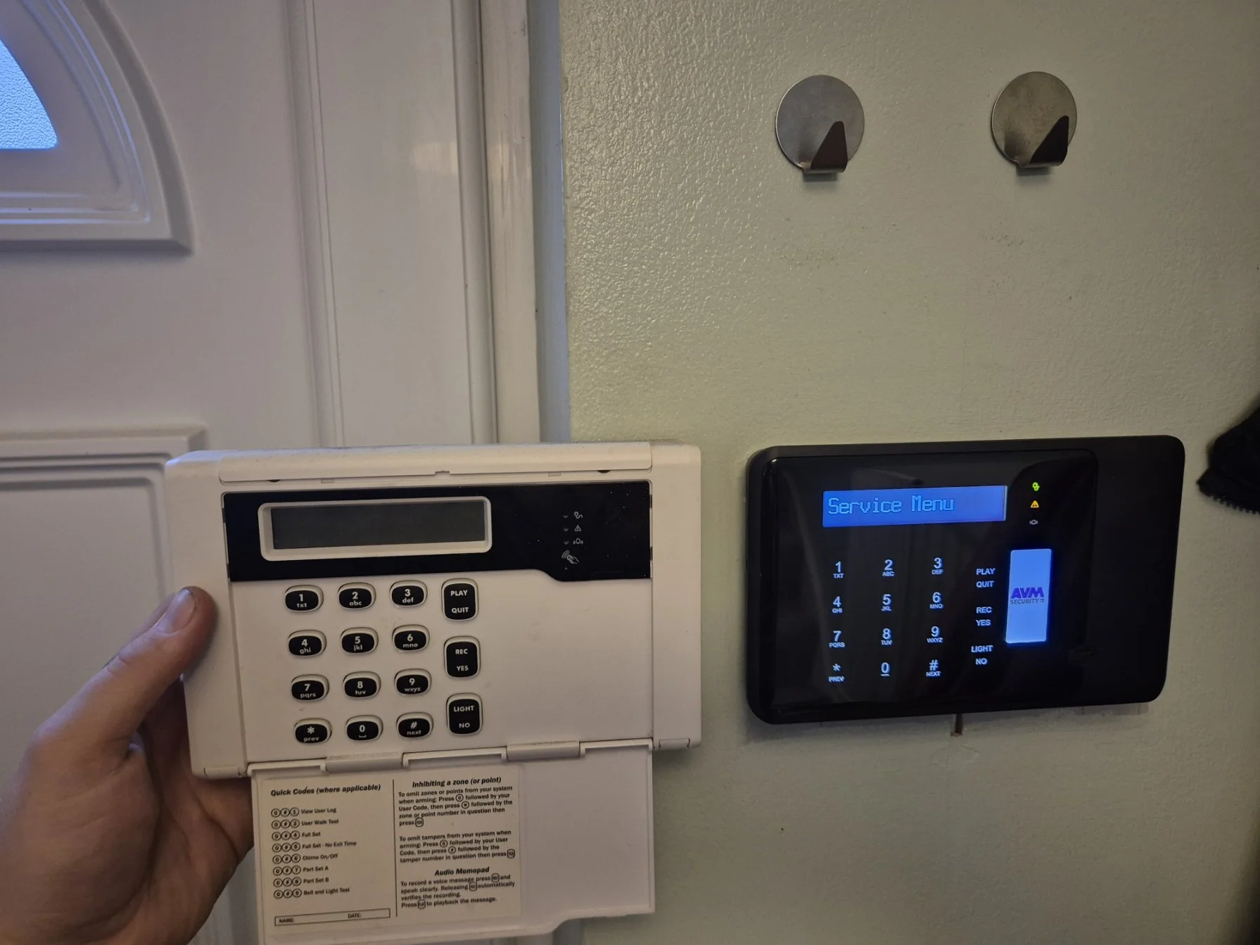 A person holding a white security keypad with a numeric keypad and function buttons, mounted on a wall near a door with an electronic security panel displaying a blue screen.