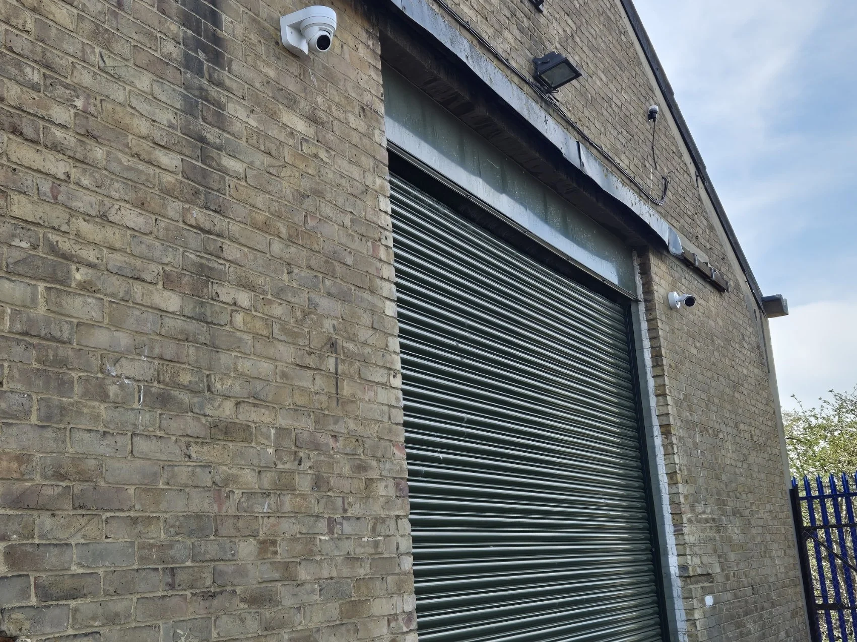 Exterior of a brick building with two security cameras mounted on the wall near a large metal roll-up garage door. The building has a weathered, industrial appearance.