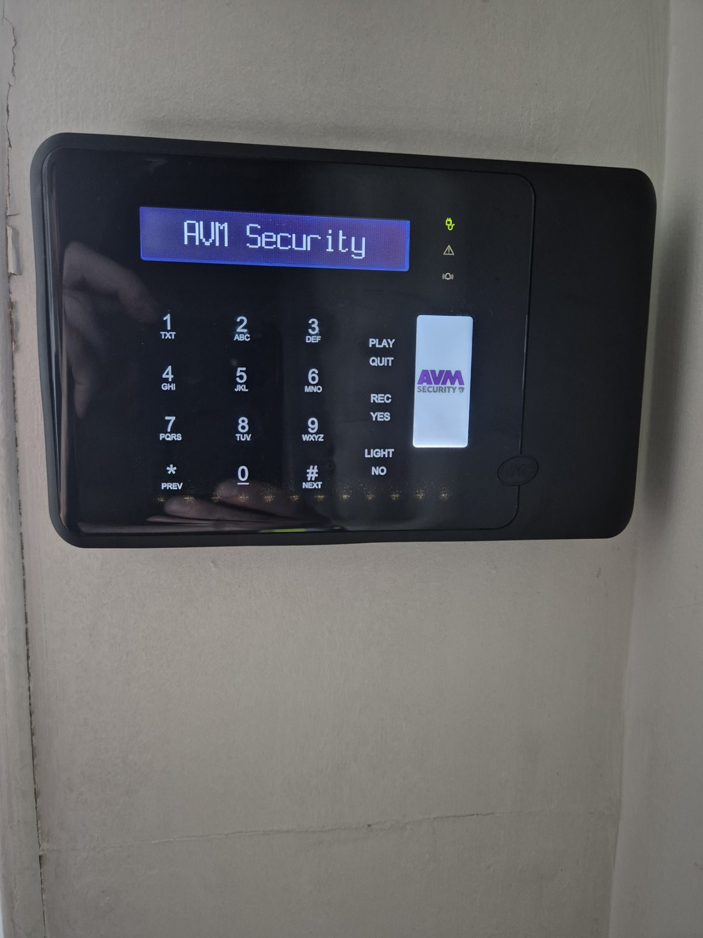 Security keypad mounted on a wall displaying 'AVM Security' with a blue backlit screen and buttons for 0-9, *, #, and functions including play, quit, record, light, and yes.