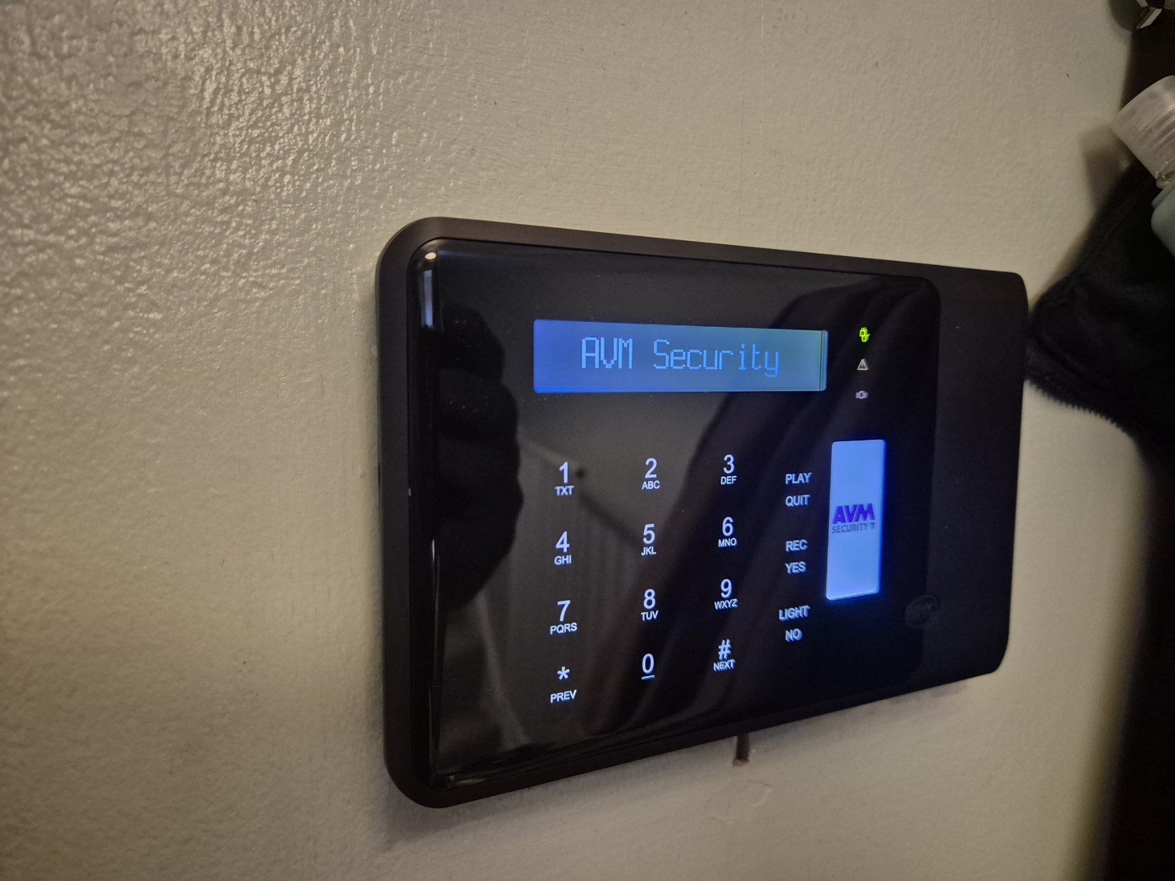 Security alarm keypad on a wall displaying 'AVM Security' on the screen and illuminated blue buttons.