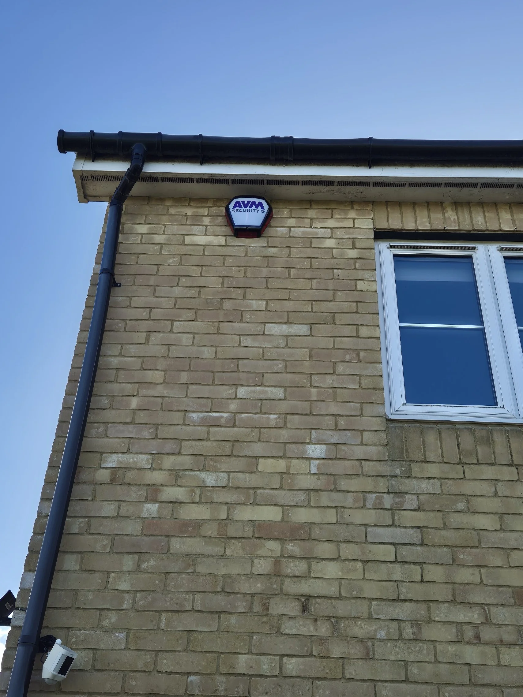 Close-up of a brick house exterior showing a security alarm device on the wall, a black gutter along the roof, a window with white frame, and a blue sky background.