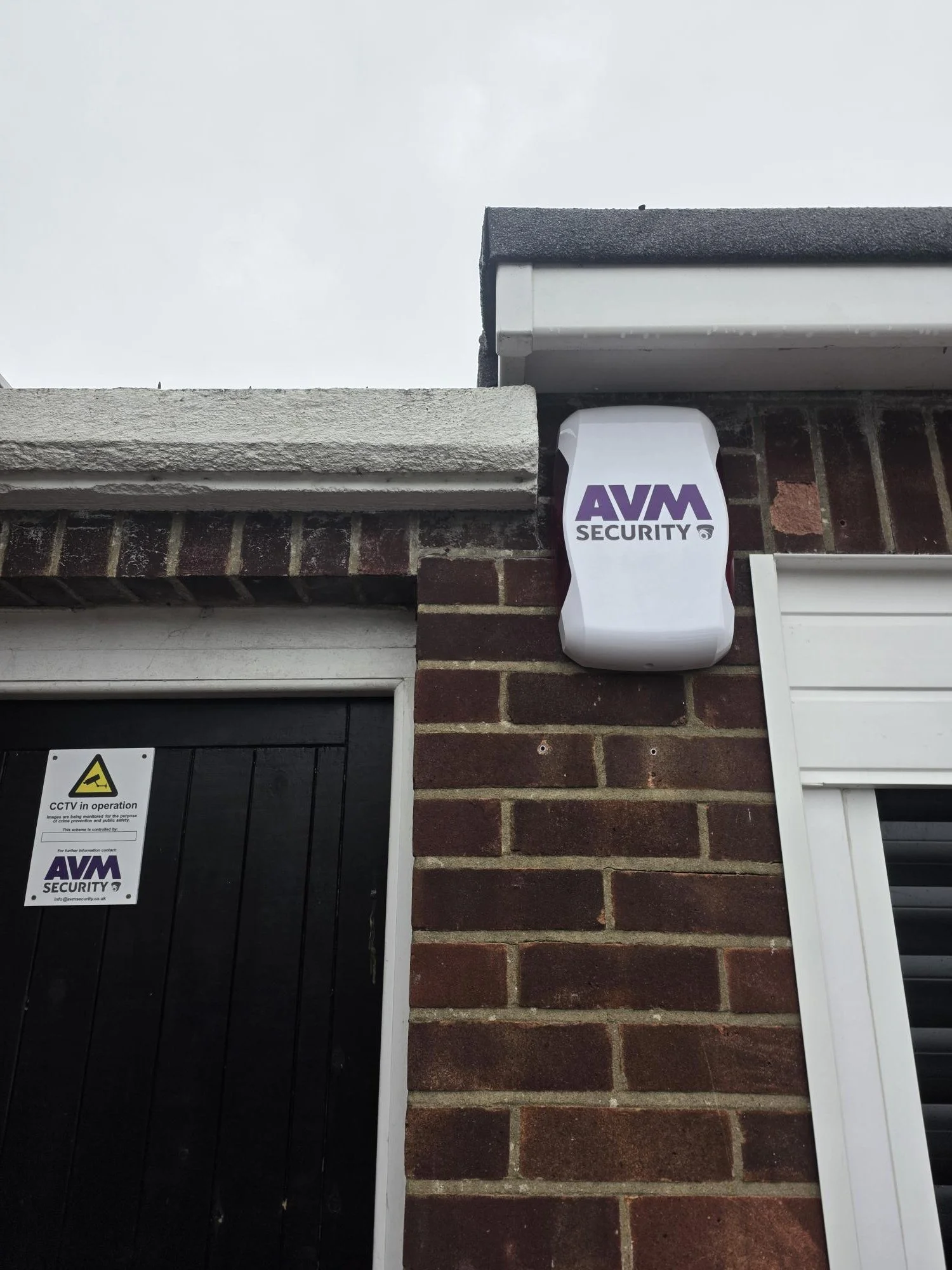 Security camera mounted on a brick building with AVM Security branding, next to a black door with a CCTV notice.