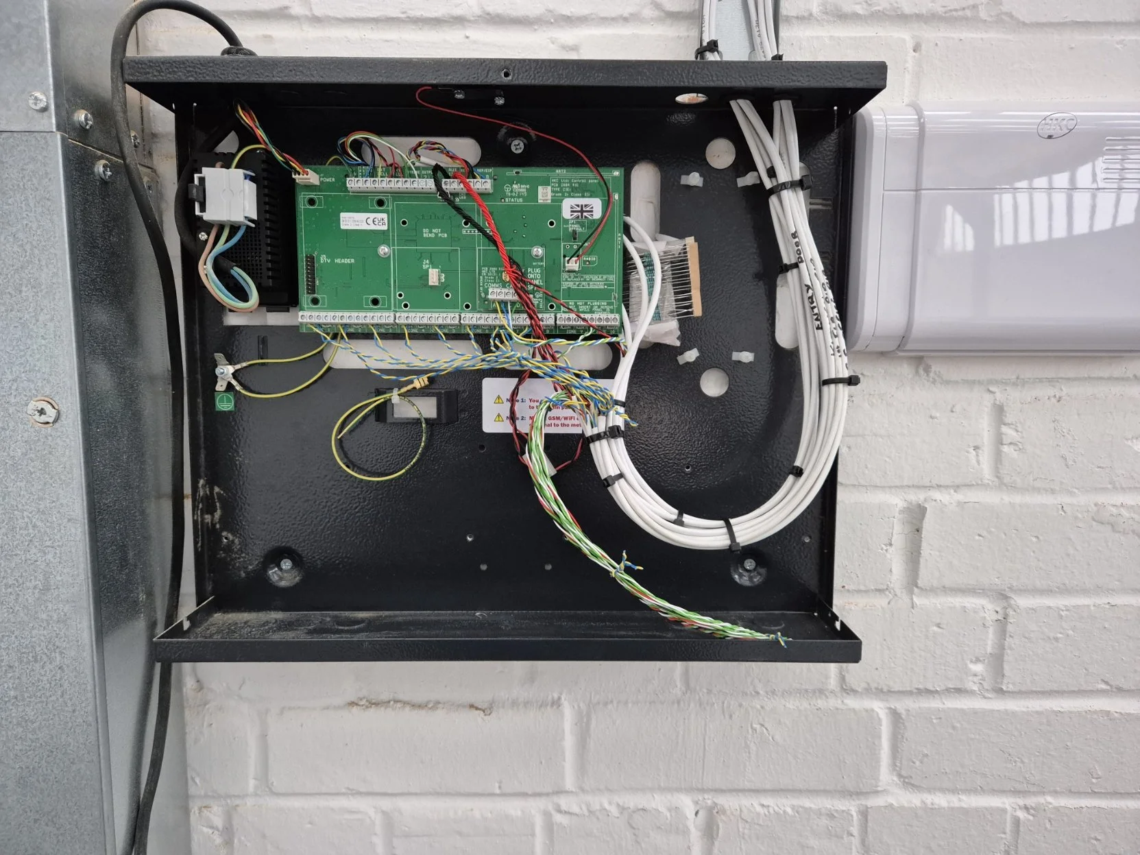 Open electrical box with circuit board, wiring, and white cables mounted on a white brick wall.