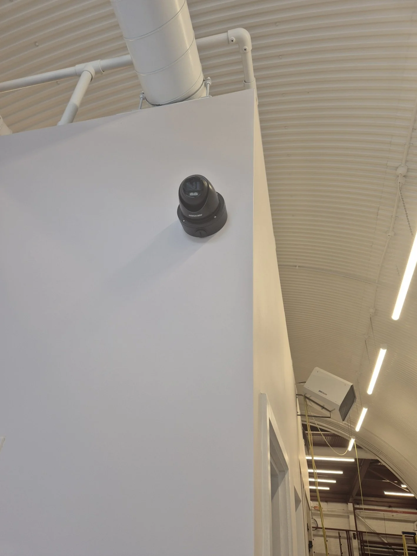 Interior of a building with a white wall, a security camera mounted on the upper part of the wall, exposed pipes, and ceiling lights.