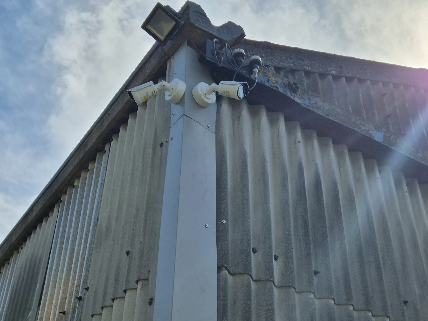 Corner of a building with surveillance cameras and a security light mounted on the wall under a cloudy sky.
