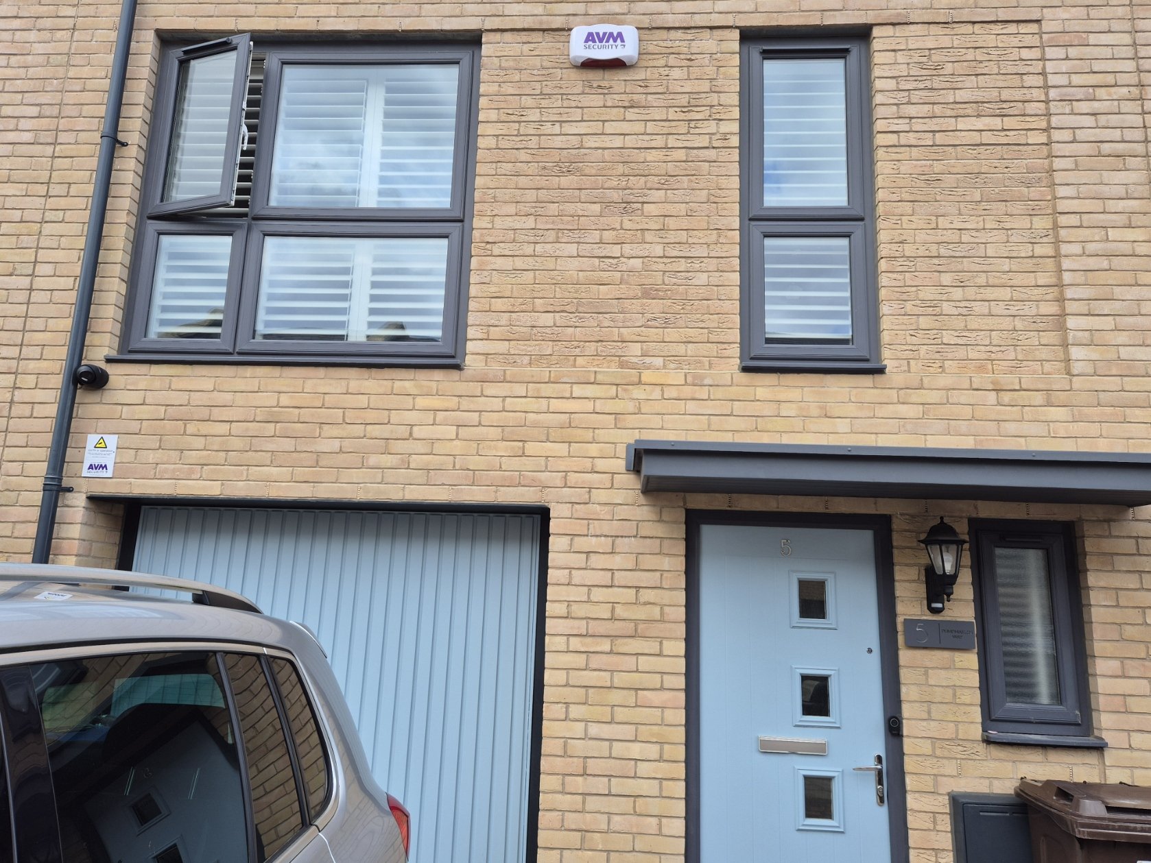 The front of a modern brick house with a light blue door that has four small square windows, a black lantern-style light fixture next to it, and a gray mailbox beneath a small window. There is a large garage door to the left, a partial view of a park