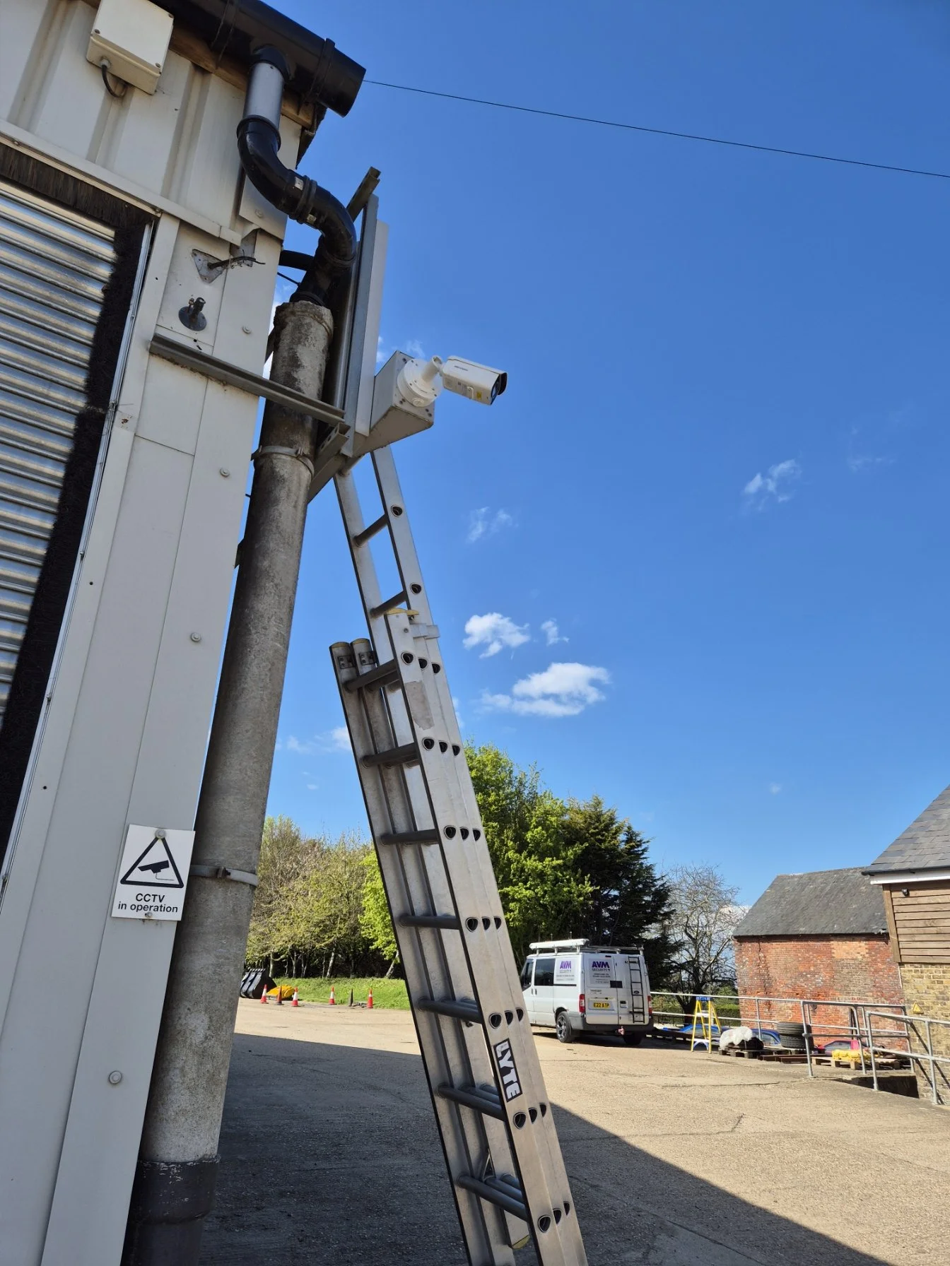 Ladder leaning against a pole beside a building with CCTV camera and utility pipes, with a van and a brick building in the background under a clear blue sky.