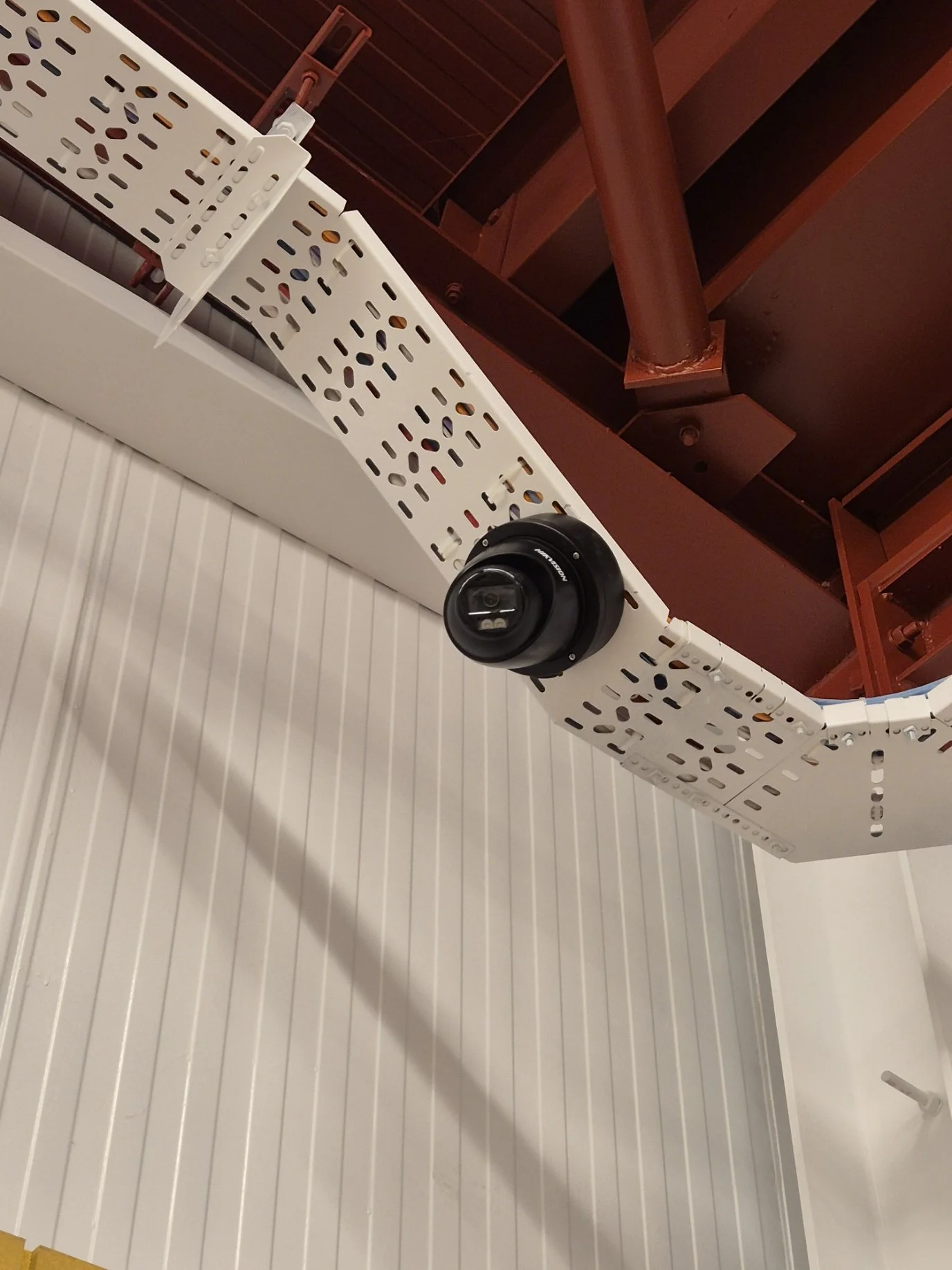 Ceiling structure with a black security camera mounted on a white, vented panel, with red-painted pipes and beams overhead.