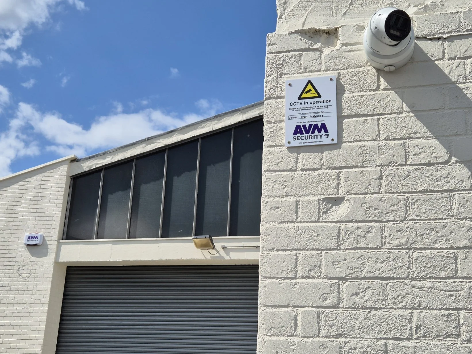 Exterior corner of a building showing a security camera, security warning signs, and a roller shutter door with the sky in the background.