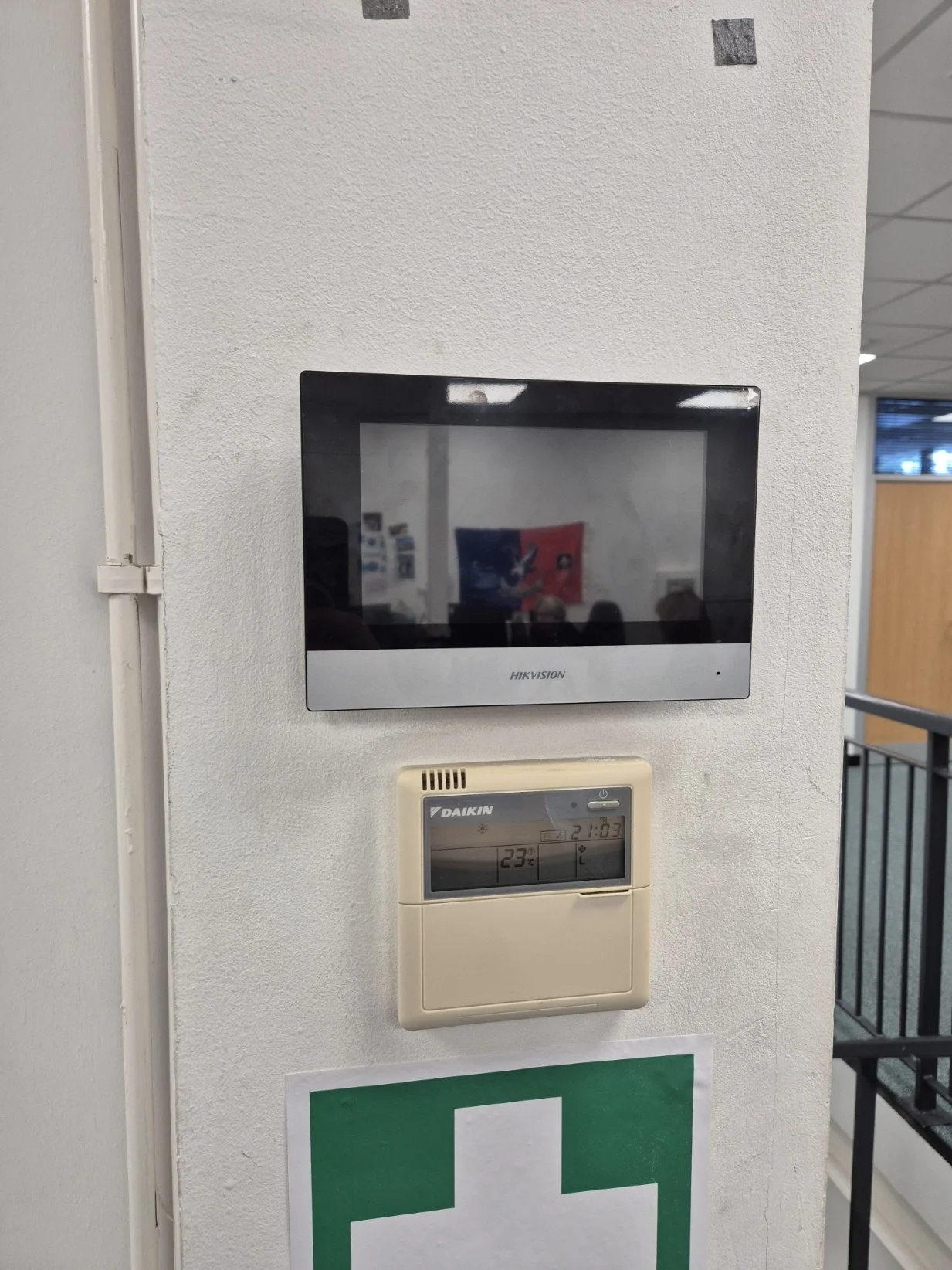 A wall with a black Hikvision security monitor, a beige Daikin thermostat showing 23°C, and a green first aid sign.