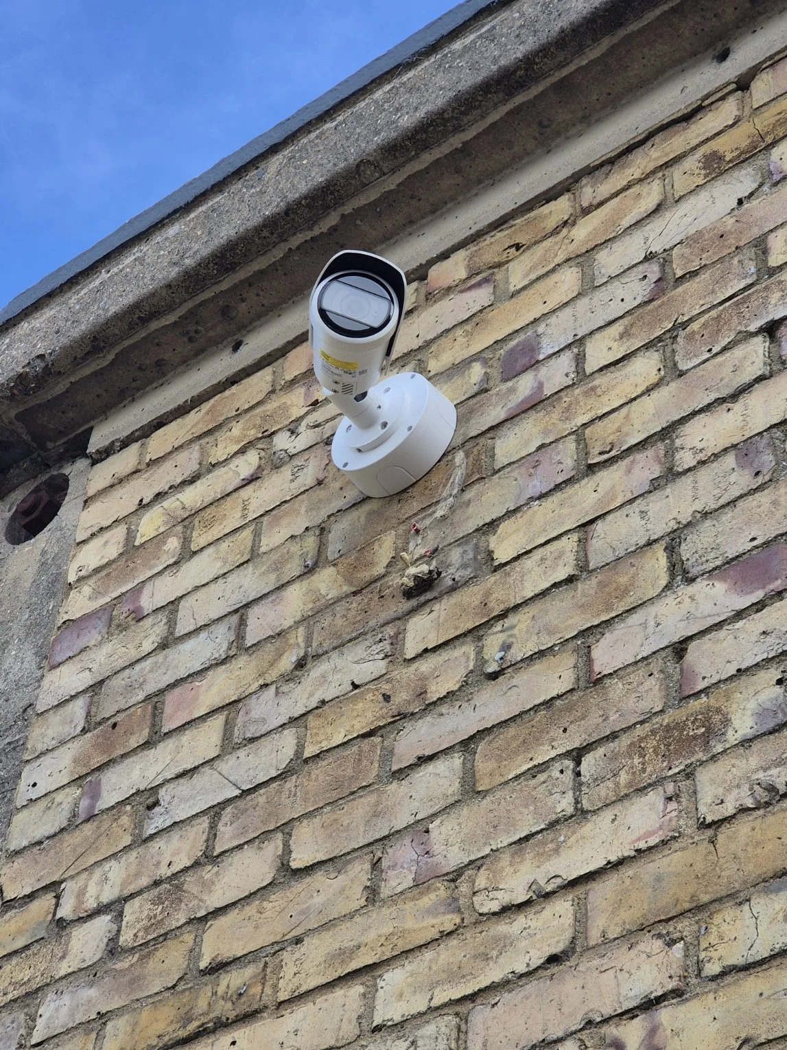 Security camera mounted on a brick wall beneath a concrete roof edge under a blue sky.