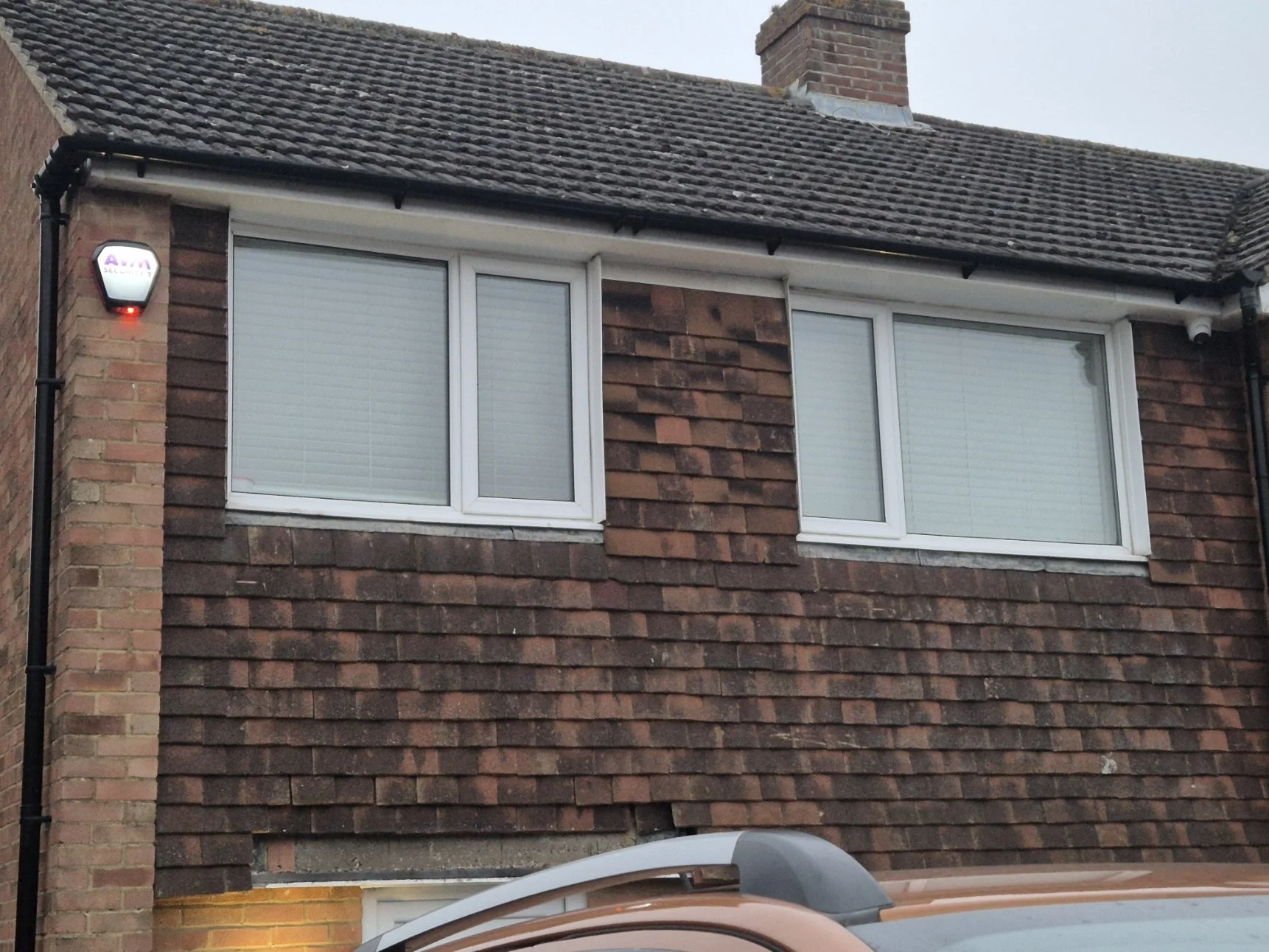 The exterior of a brick house with two white-framed windows on the upper floor, a red and white security alarm on the left, a brick chimney, a sloped roof with brown tiles, and part of a gray vehicle in the foreground.
