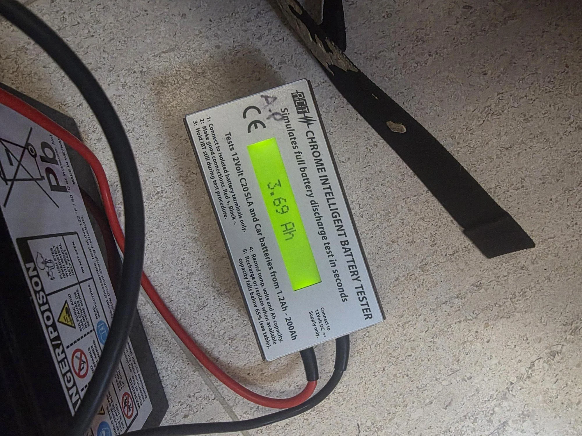 A digital battery tester displaying 3.69 volts, connected to an alarm back-up battery with red and black wires. The tester is placed on a surface near the battery.