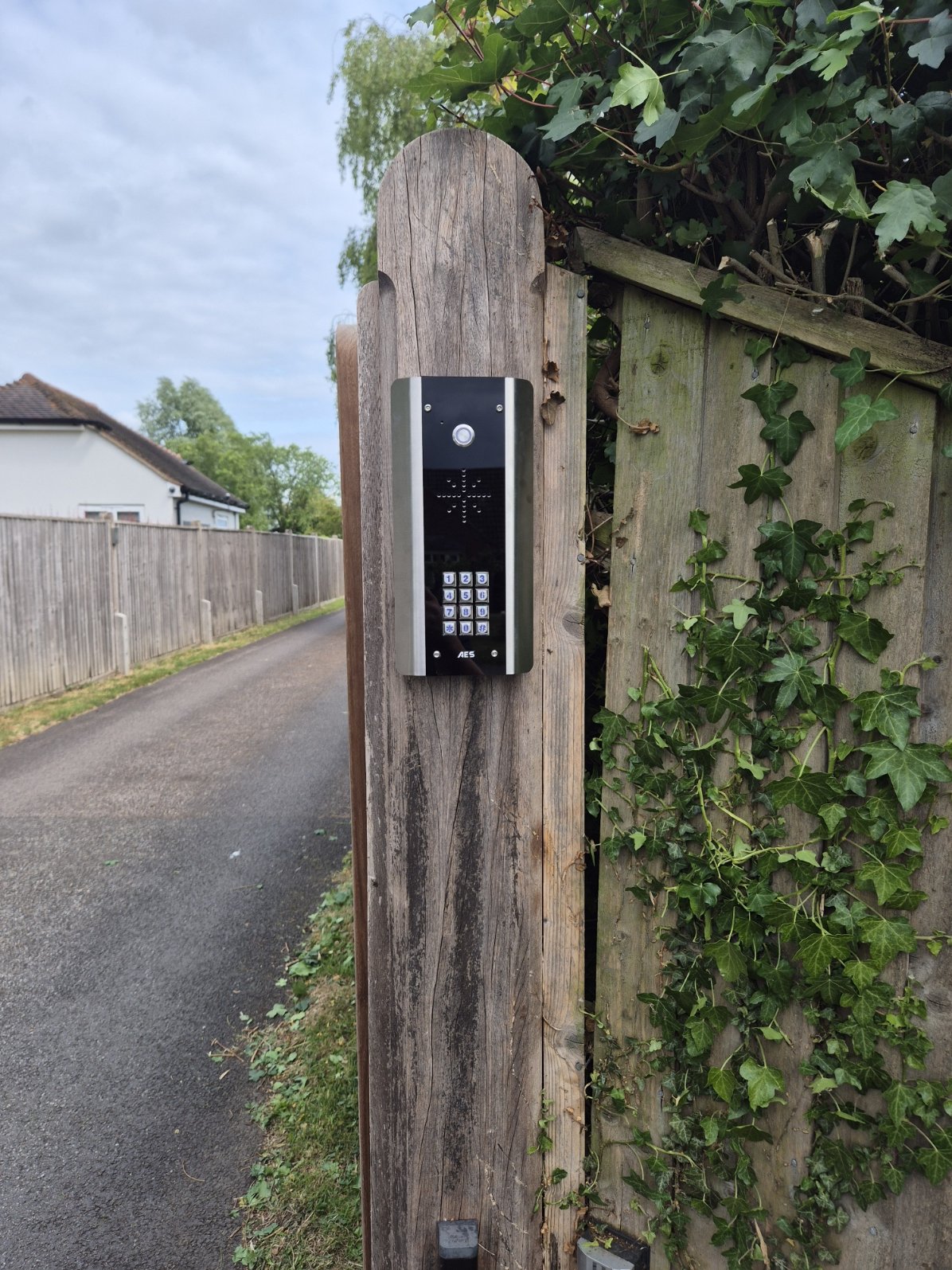 Intercom with keypad attached to a wooden fence post beside a garden fence, with a residential street extending into the distance and cloudy sky above.
