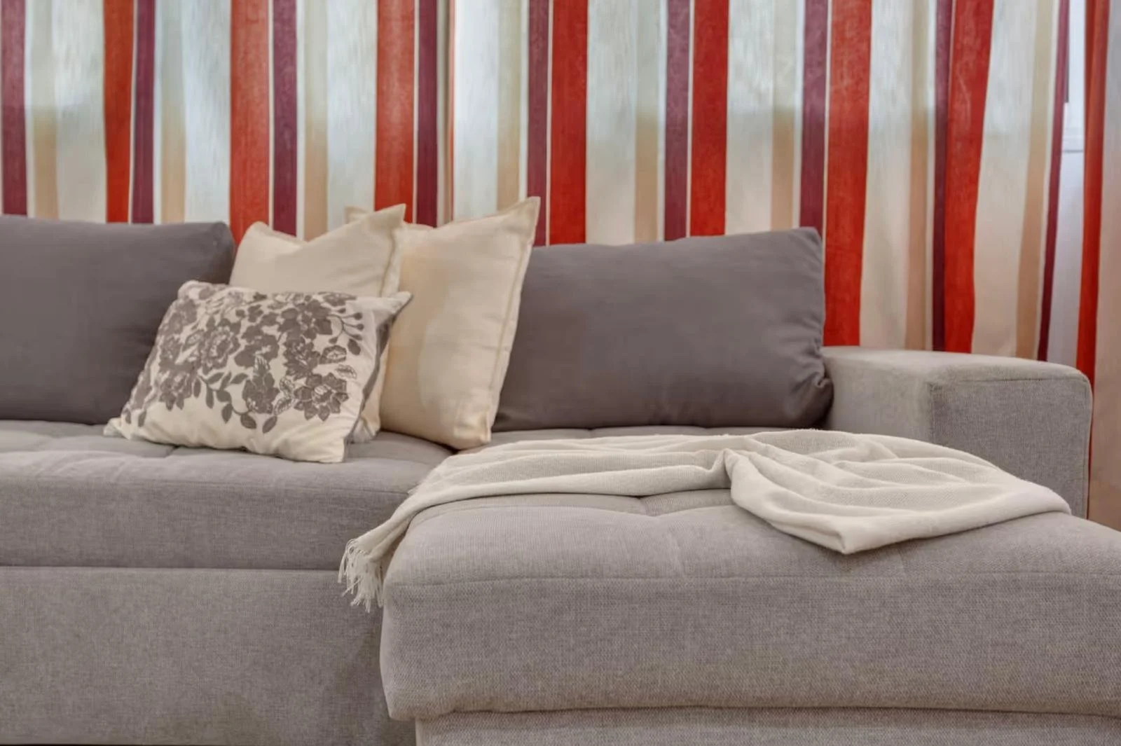 Sofá gris con varias almohadas blancas y grises y una manta blanca enrollada, en un fondo con pared de rayas rojas, beige y blancas.