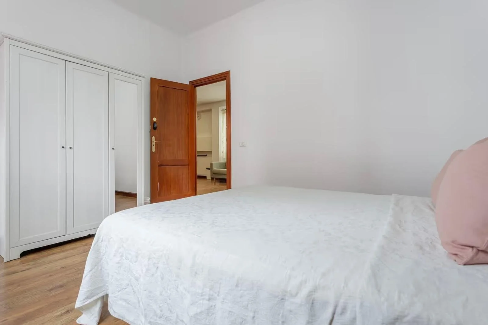 Cuarto con cama, ropa de cama blanca, almohada rosa, armario blanco y puerta de madera, piso de madera, paredes blancas.