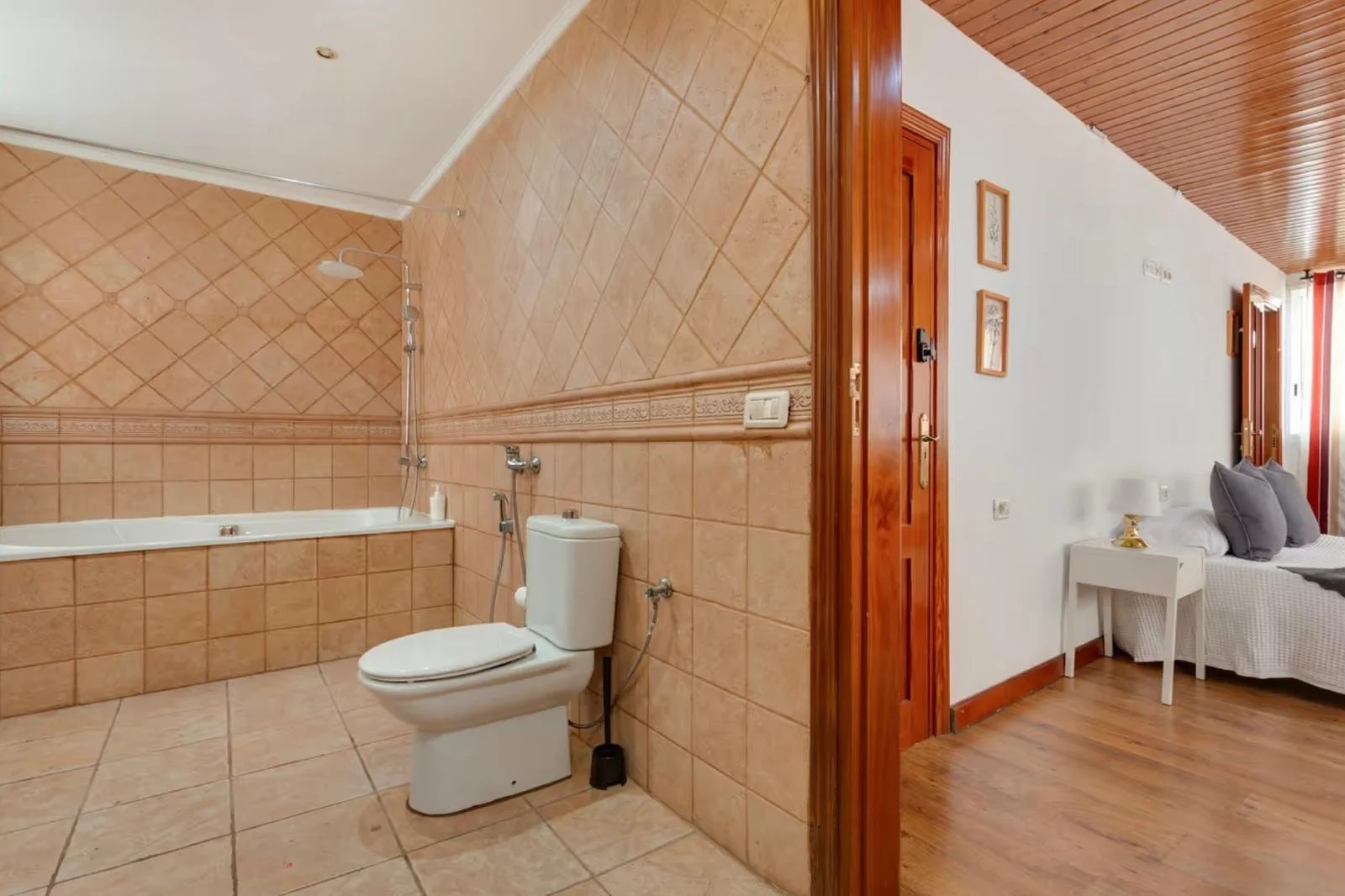 Baño con azulejos color terracota, bañera, inodoro y accesorios de baño. A la derecha, vista de una habitación con piso de madera, sofá y lámpara de mesita de noche.