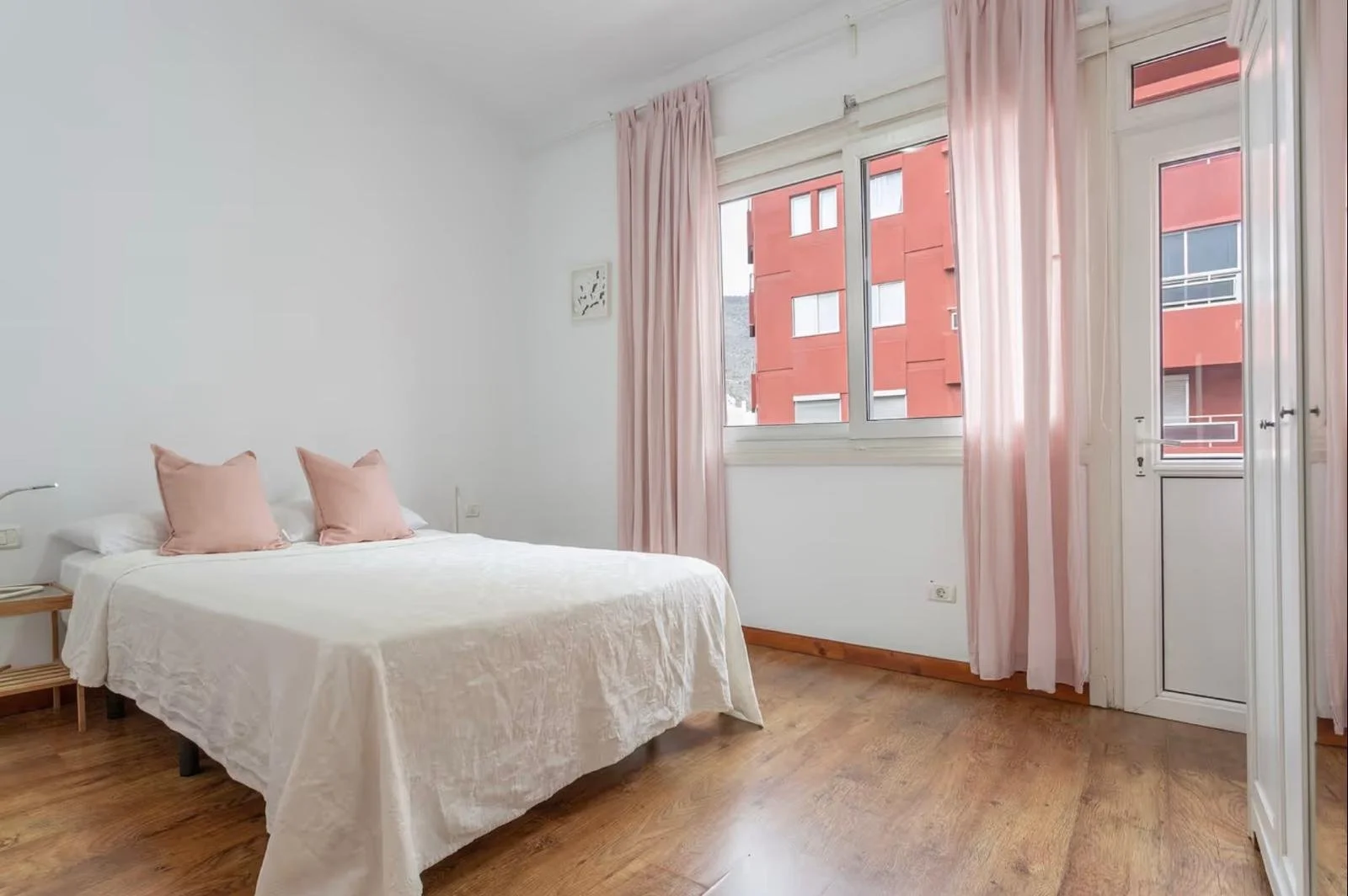 Habitación con cama doble, cortinas rosadas, ventanas grandes con vista a edificio rojo.