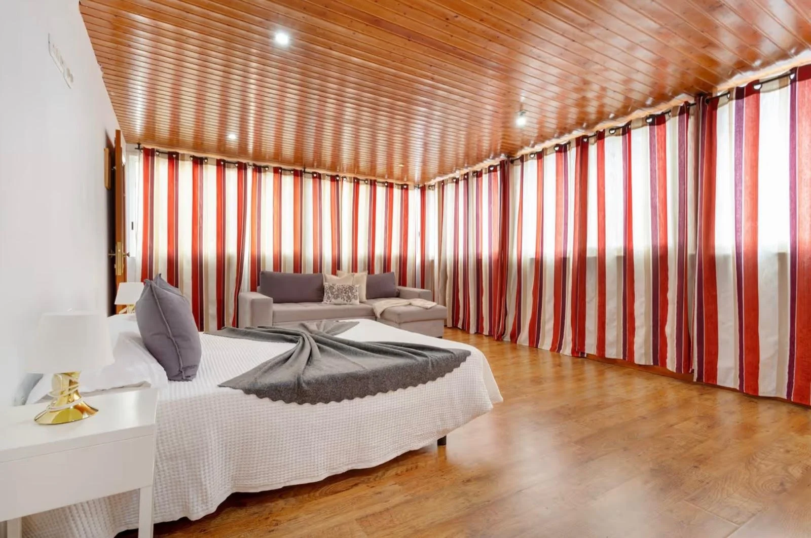 Habitación con cama, sofá gris, cortinas de rayas rojas y blancas, suelo de madera y techo de madera