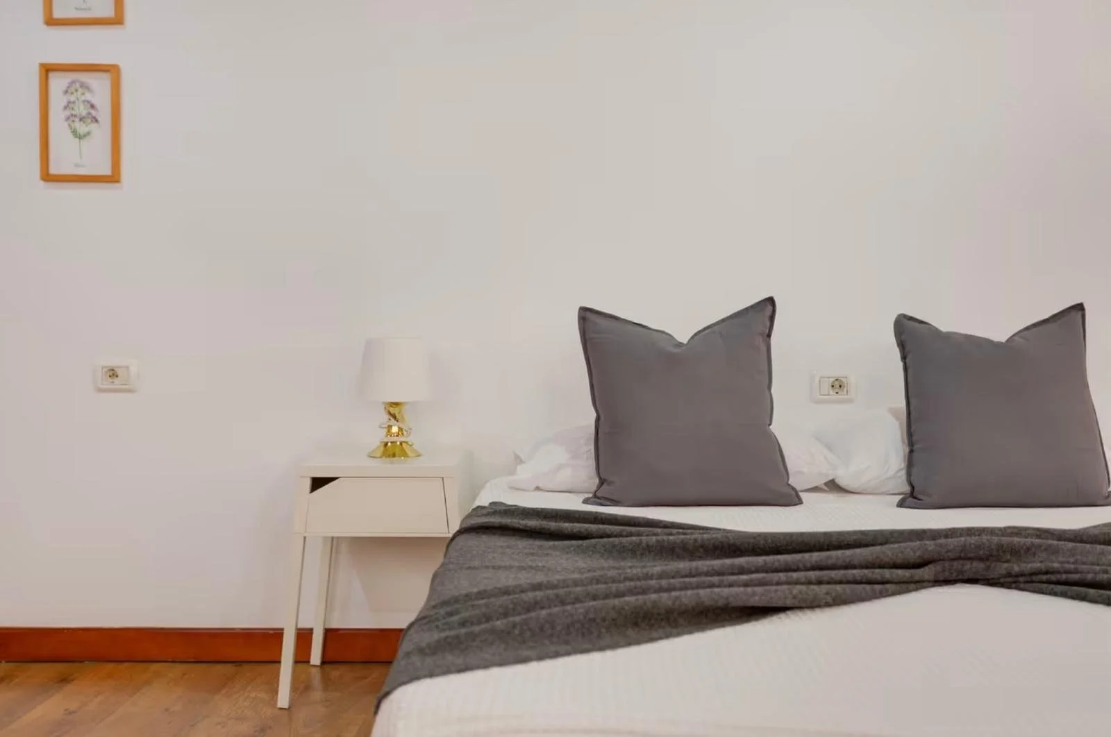 Cama con dos cojines grises, una lámpara dorada y una mesita blanca junto a la cama, decoraciones en la pared.