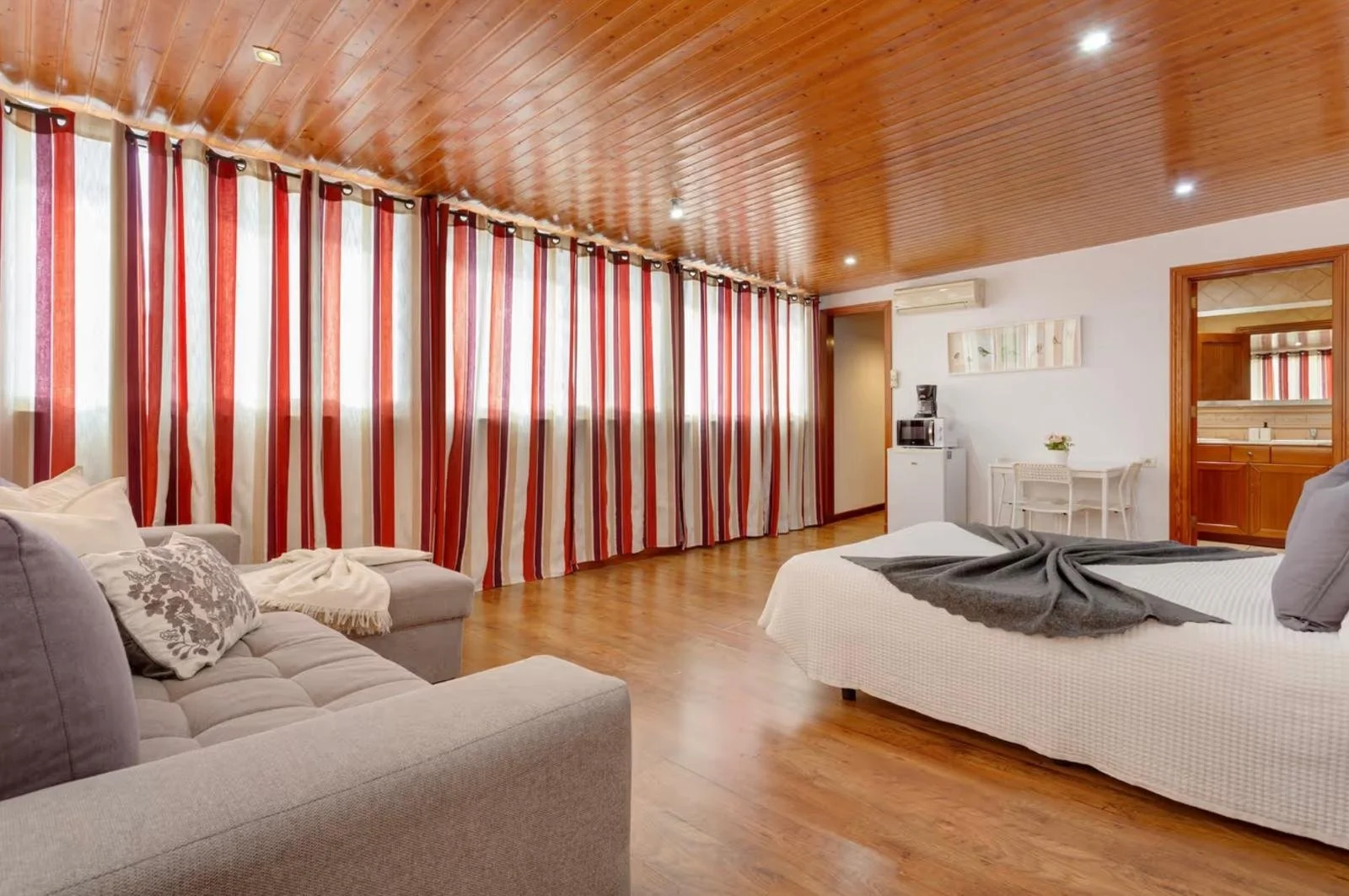Habitación de hotel con paredes de madera, cortinas a rayas rojas, blancas y beige, sofá gris con cojines, cama con ropa blanca y manta gris, suelo de madera, y área de kitchenette con microondas y mesa blanca.