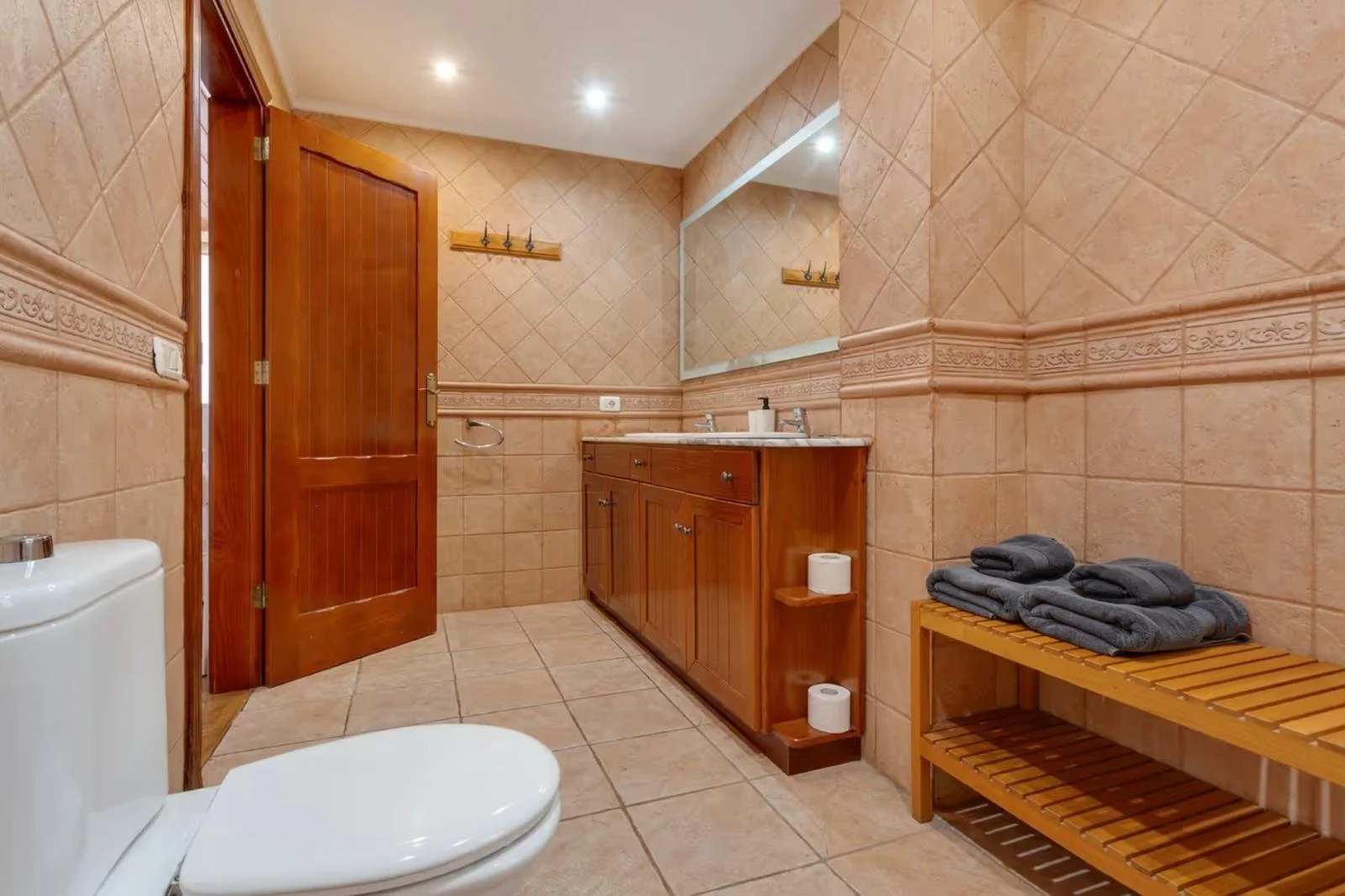 Baño con paredes y piso de azulejos beige, lavabo de madera con espejo grande, toallas enrolladas sobre un banco de madera, y un inodoro blanco.