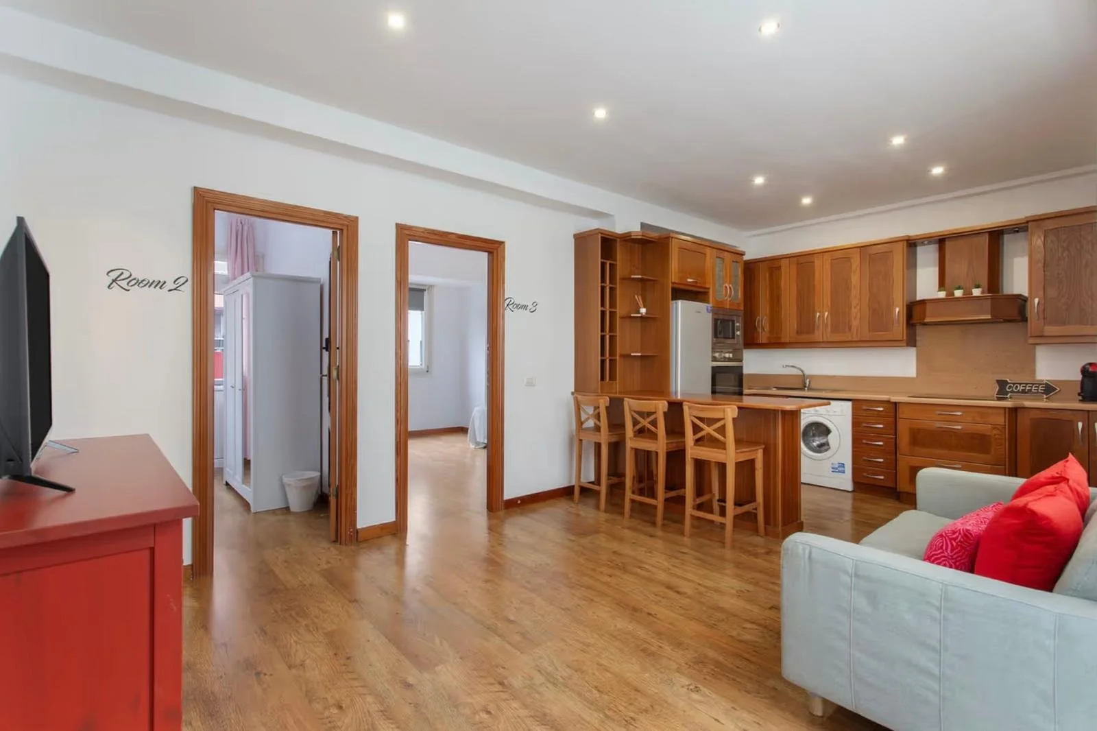 Vivienda moderna con cocina y sala de estar, paredes blancas, suelo de madera, puertas de madera, con sillón gris y cojines rojos, y recámaras visibles a través de las puertas.