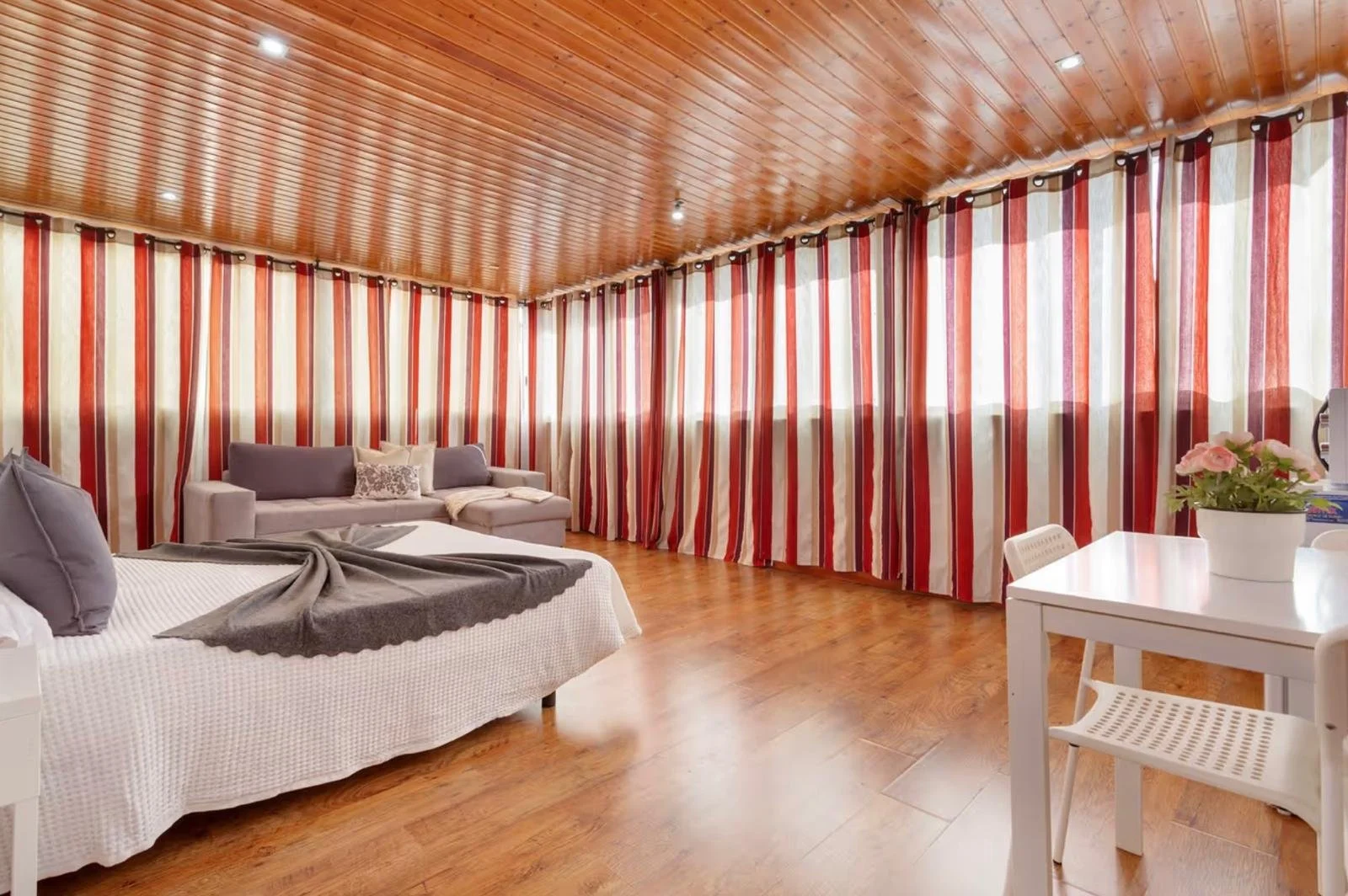 Habitación con paredes y cortinas de rayas rojas, blancas y beige, suelo de madera, cama con sábanas blancas y almohadas grises, sofá gris con cojín decorativo, mesa blanca con una planta y una máquina de café.