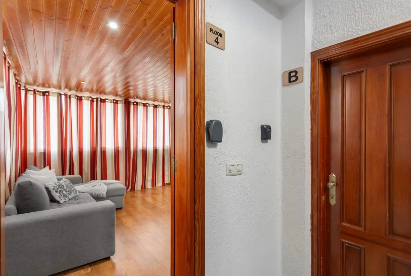 Vista del pasillo de un apartamento o casa con puerta de madera, señal que indica piso 4 y letra B, y una sala con cortinas de rayas en fondo rojo, beige y blanco, sillón gris, cojines y una manta en el sofá, paredes blancas y piso de madera.