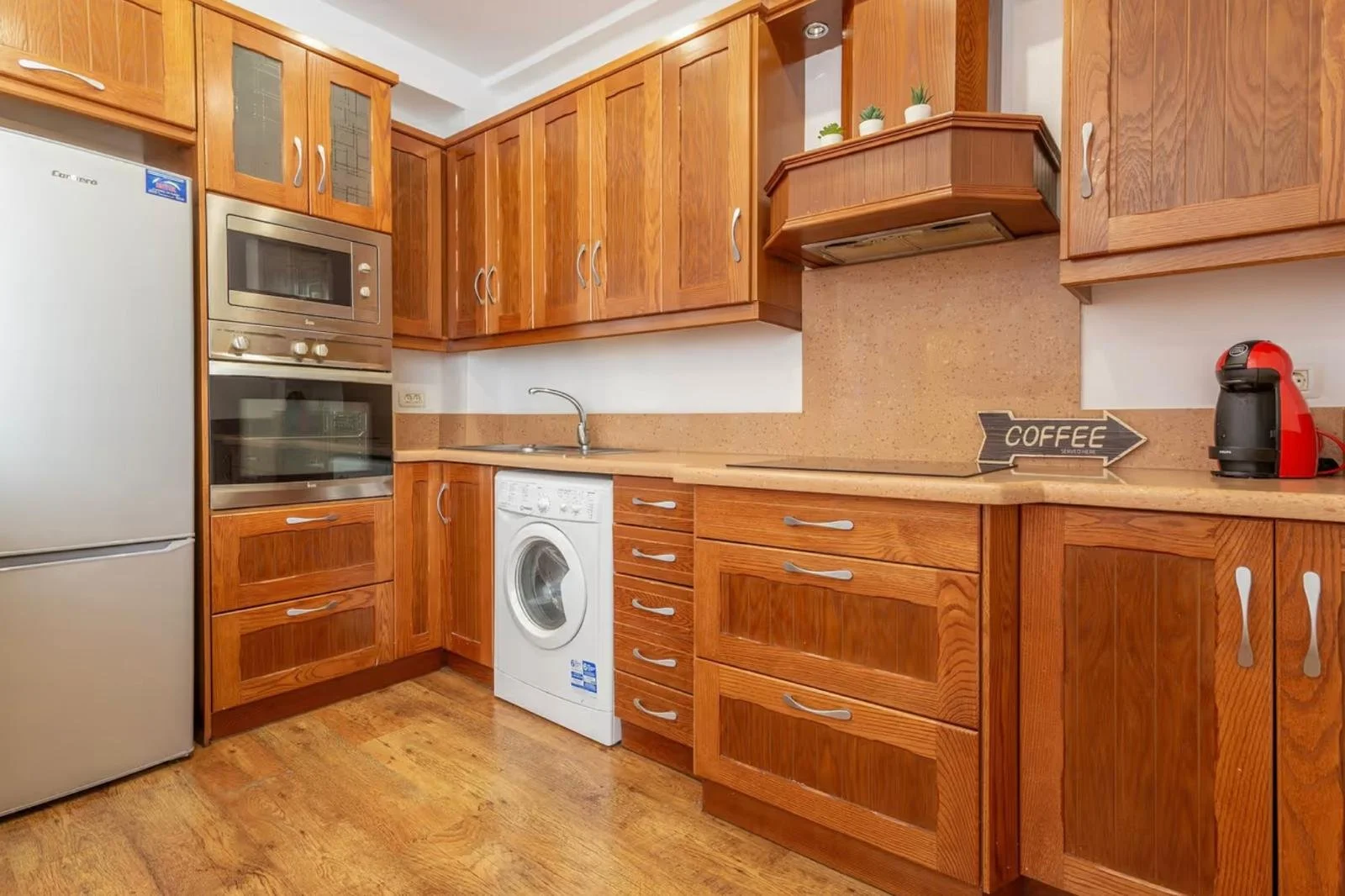 Cocina con gabinetes de madera, electrodomésticos de acero inoxidable, mesa de trabajo y señal que dice 'COFFEE' en la encimera, junto a una cafetera roja.