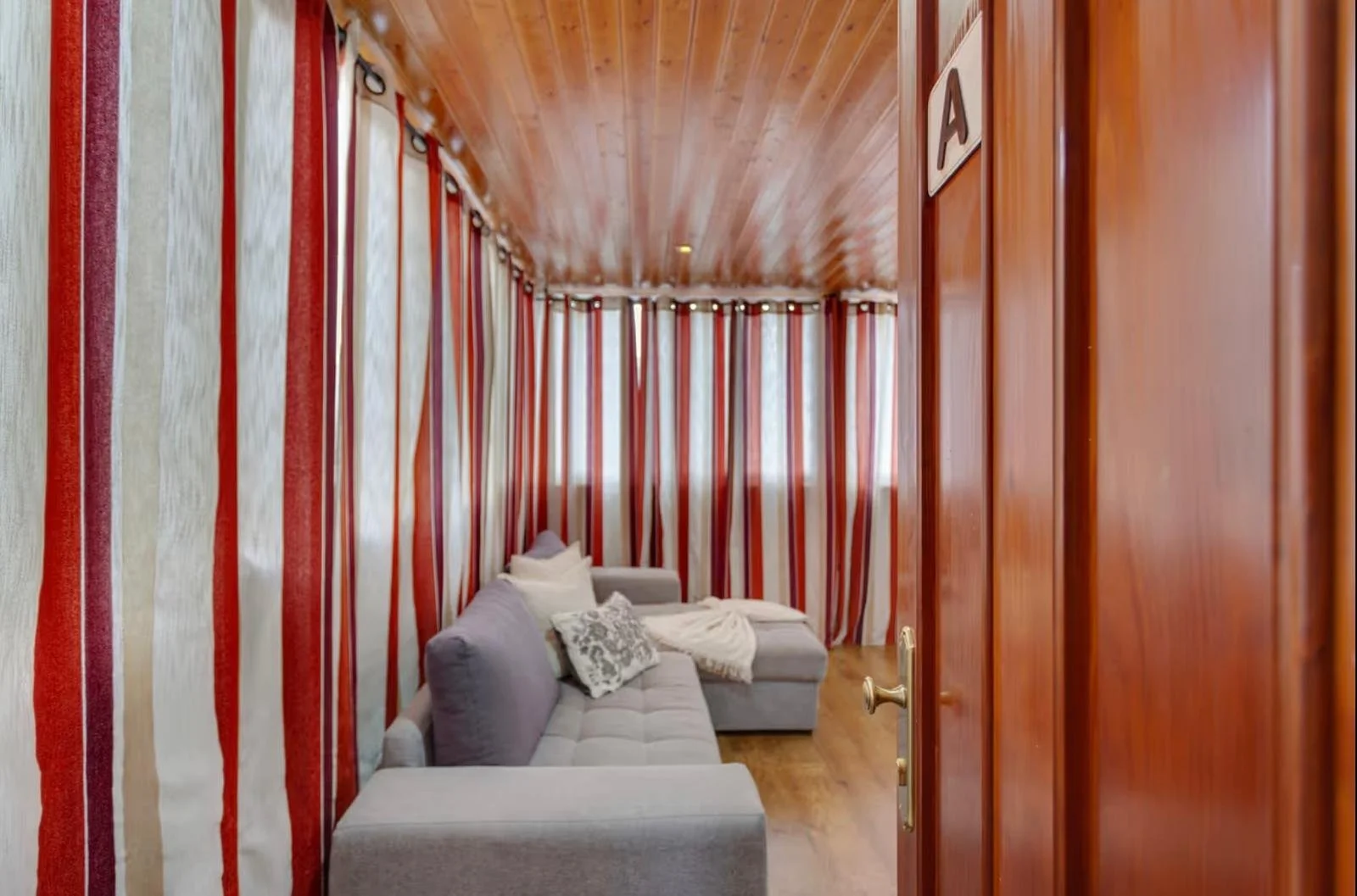 Sala de espera con cortinas a rayas en tonos rojos y beige, sofá gris con cojines blancos y decoración acogedora.