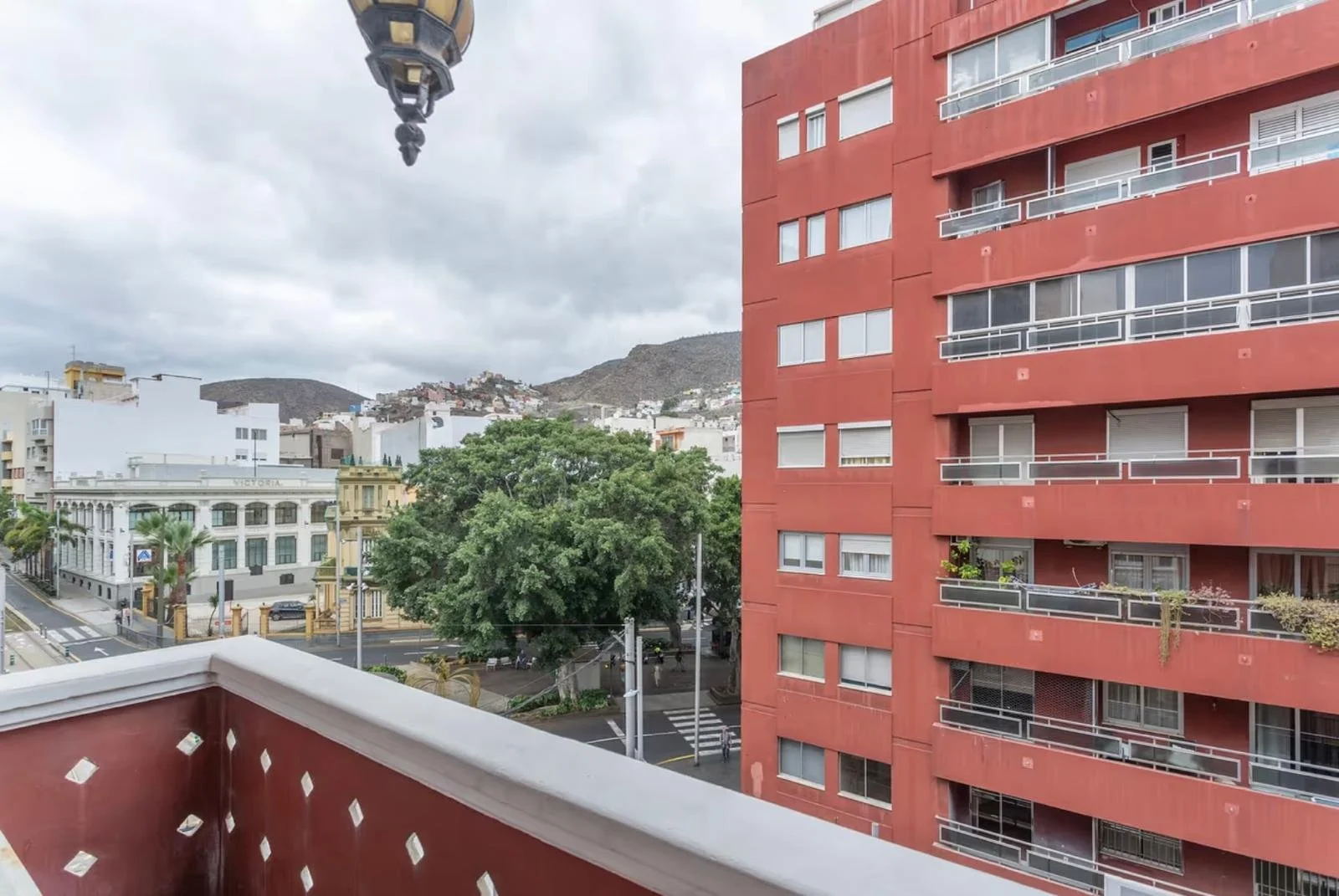 Vista desde un balcón de un edificio rojo hacia una calle con árboles y edificios, con montañas y cielo nublado al fondo.