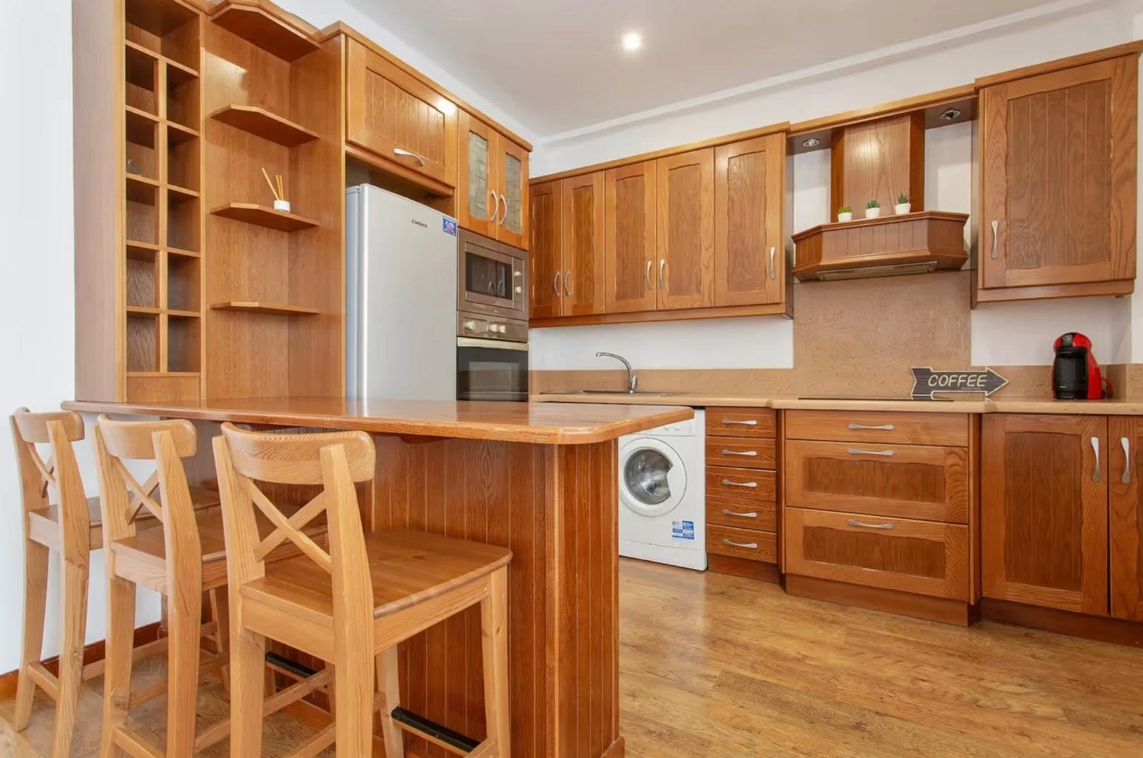 Cocina de madera con barra y cuatro sillas, electrodomésticos como refrigerador, horno, lavadora, y cafetera, con plantas pequeñas y señal de 'coffee' en la pared.