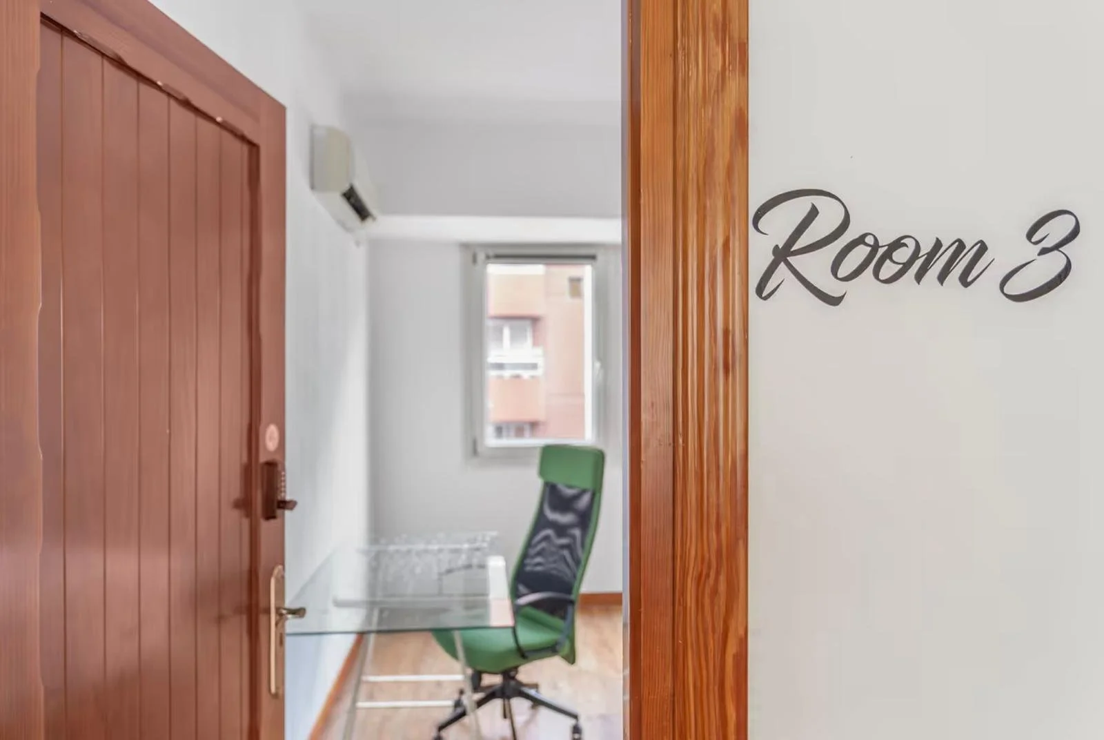 Puerta de madera y zona interior que indica 'Room 3', vista desde el pasillo con una silla verde y ventana con vista exterior.