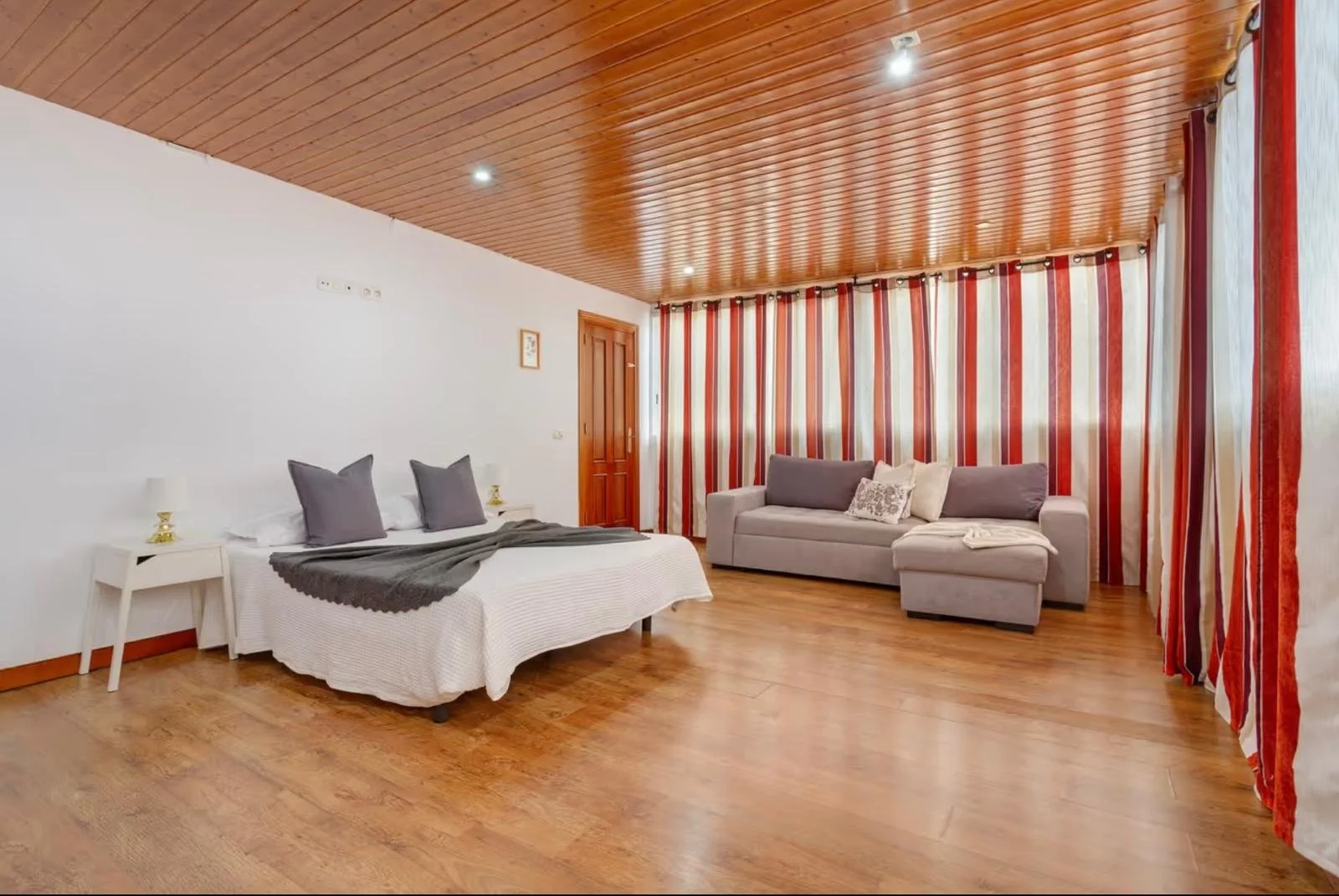 Habitat con cama, sillón, cortinas rojas y blancas, paredes blancas y techo de madera.