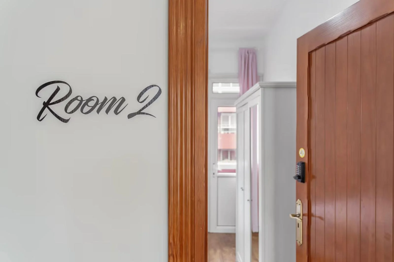 Vista del interior de una casa mostrando la entrada a la habitación marcada como "Room 2" en la pared, con una puerta de madera y detalles de mobiliario en el fondo, incluyendo una ventana con cortinas rosadas.