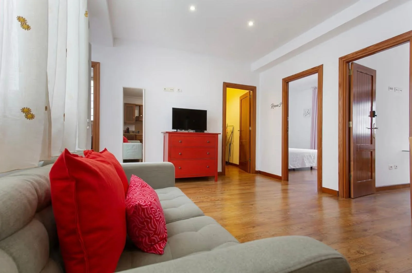 Sala con sillón beige, cojines rojos y decoraciones de cortinas con flores, madera en puertas y pisos, televisor en mueble rojo, habitaciones visibles con puertas abiertas.