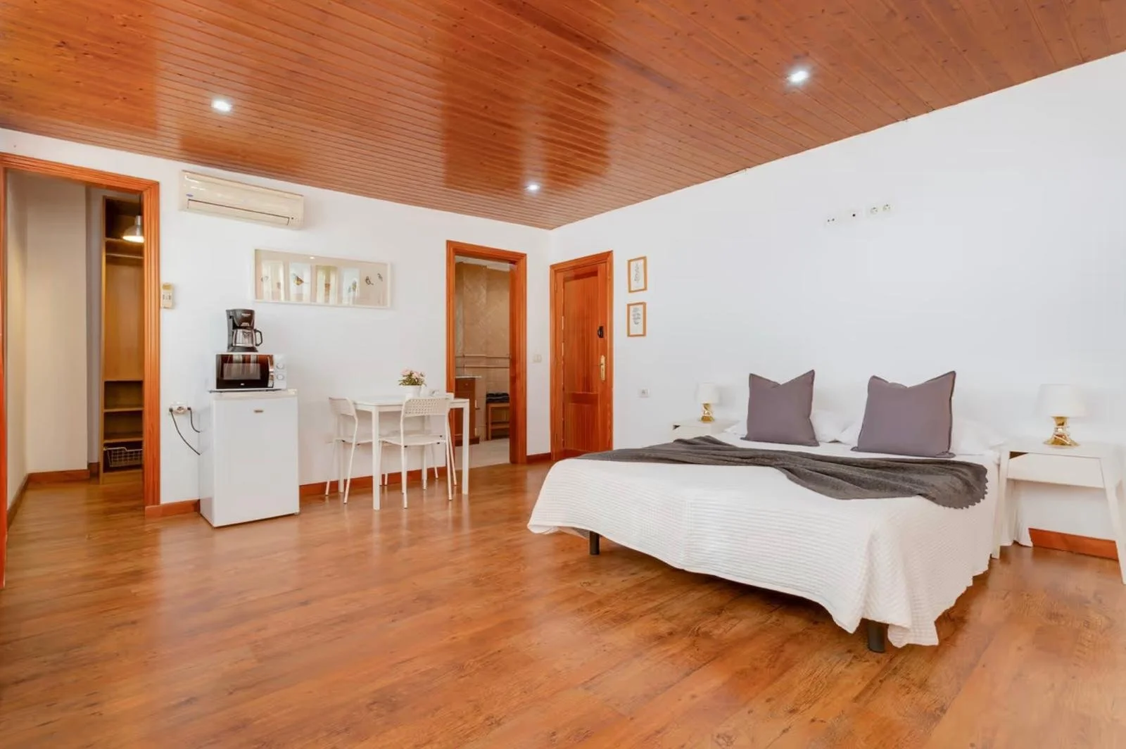 Habitación con suelo de madera, cama con sábanas blancas y almohadas grises, dos mesas de noche con lámparas, comedor con mesa y sillas, aire acondicionado, puertas de madera, decoración sencilla.