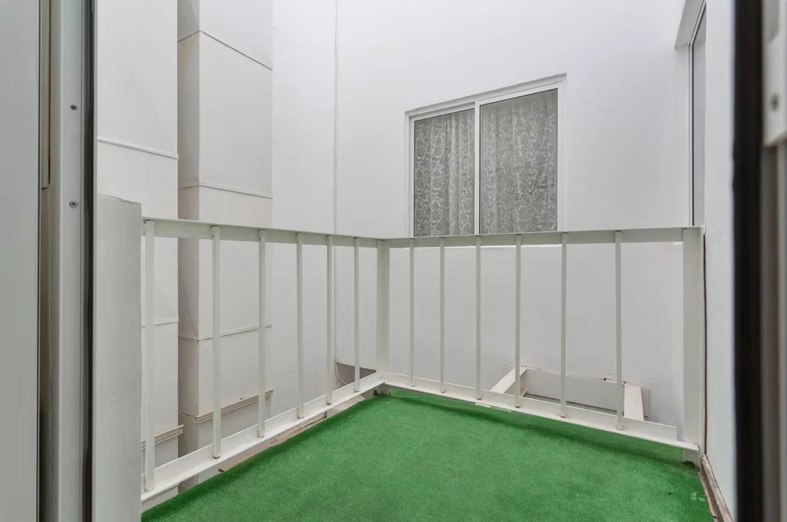 Pequeño balcón con césped artificial y barandilla blanca, en un edificio de paredes blancas y ventana con cortina de encaje.