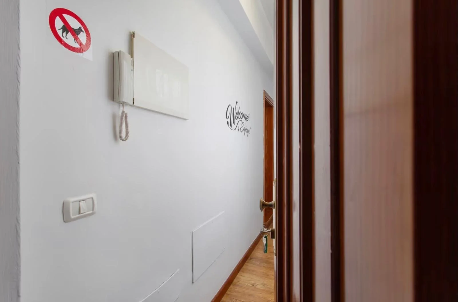 Pasillo con pared blanca, señal de prohibido perros, teléfono en la pared, pared con escritura que dice 'Welcome' en inglés, puertas de madera cerradas