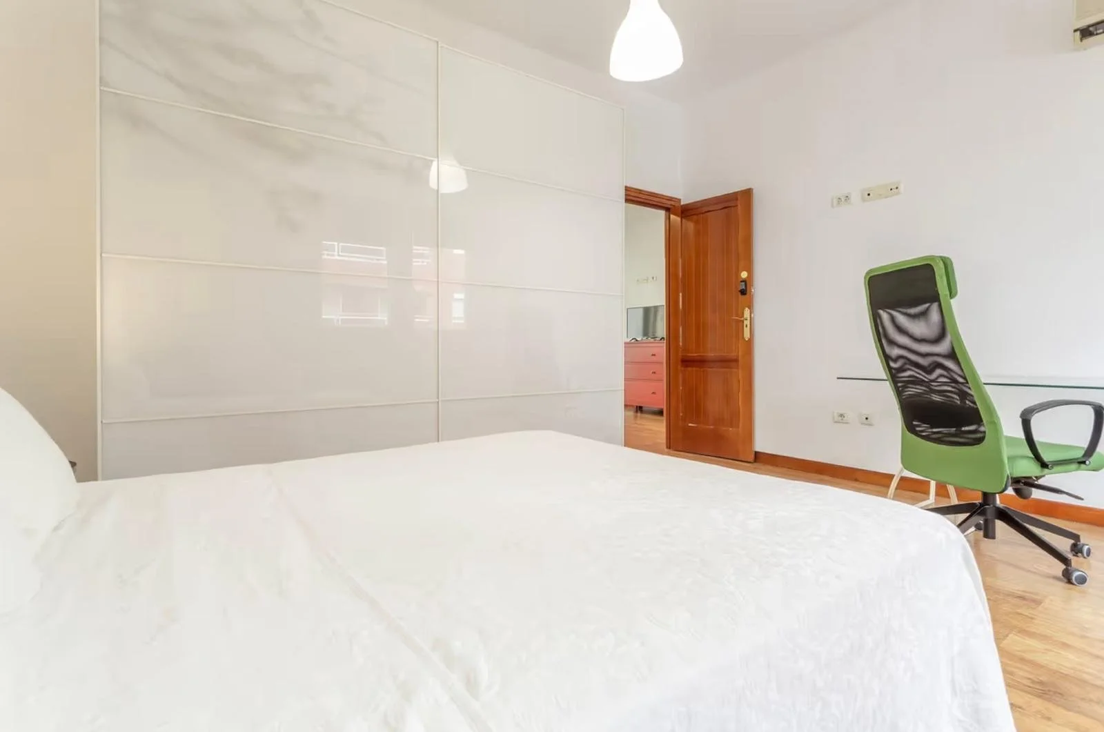 Dormitorio con cama blanca, armario grande blanco, puerta de madera, silla de oficina verde con respaldo negro y escritorio minimalista.
