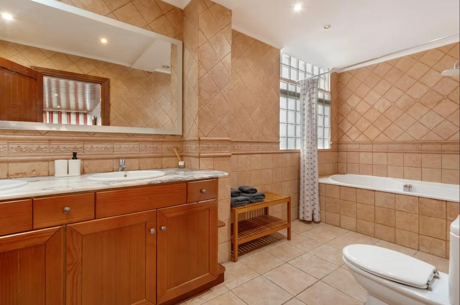 Baño amplio con lavabo de madera, espejo grande, ducha y bañera con cortina, y ventanas con bloques de vidrio.