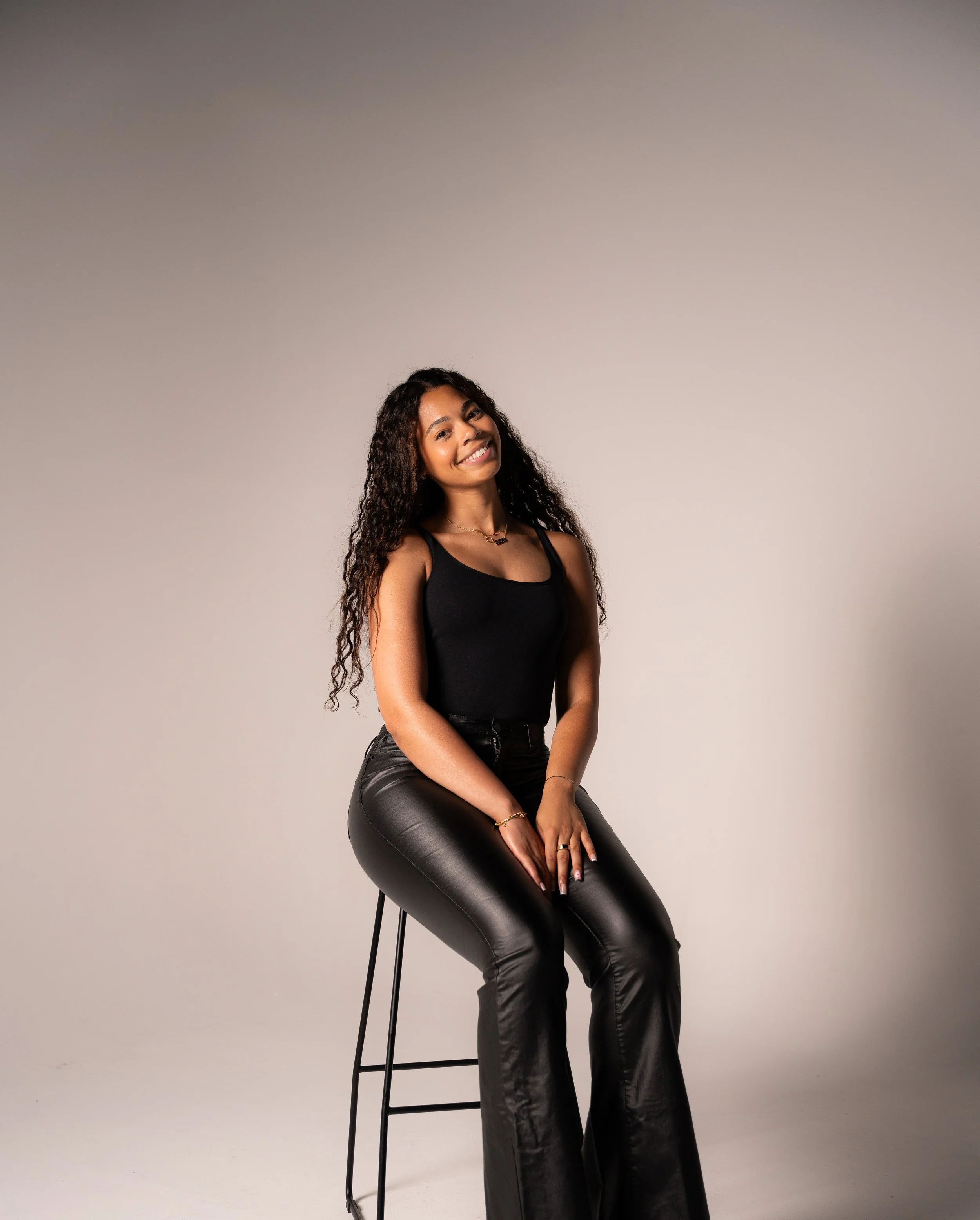A woman with long curly hair is sitting on a tall black stool against a plain light gray background. She is wearing a black tank top and black leather pants, smiling at the camera.