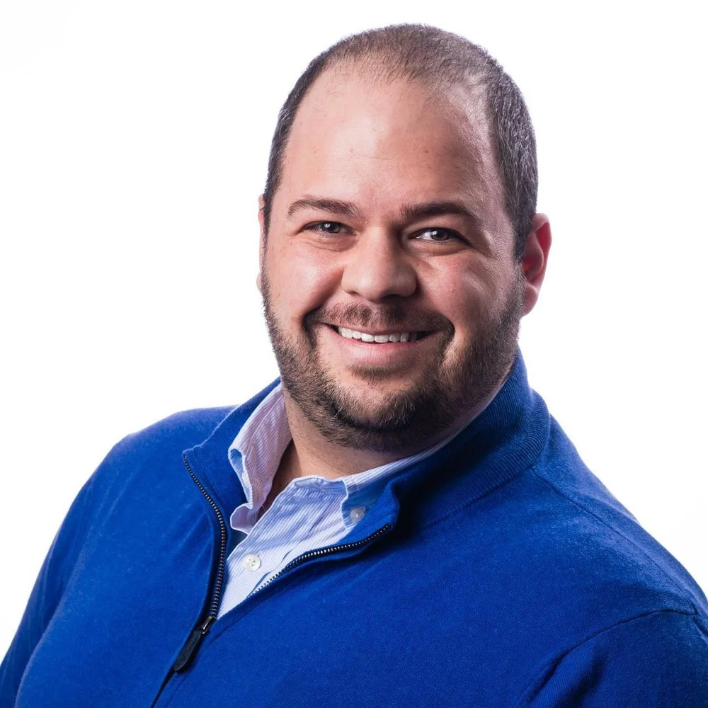 A smiling man with a beard and short hair wearing a blue jacket and a white collared shirt.
