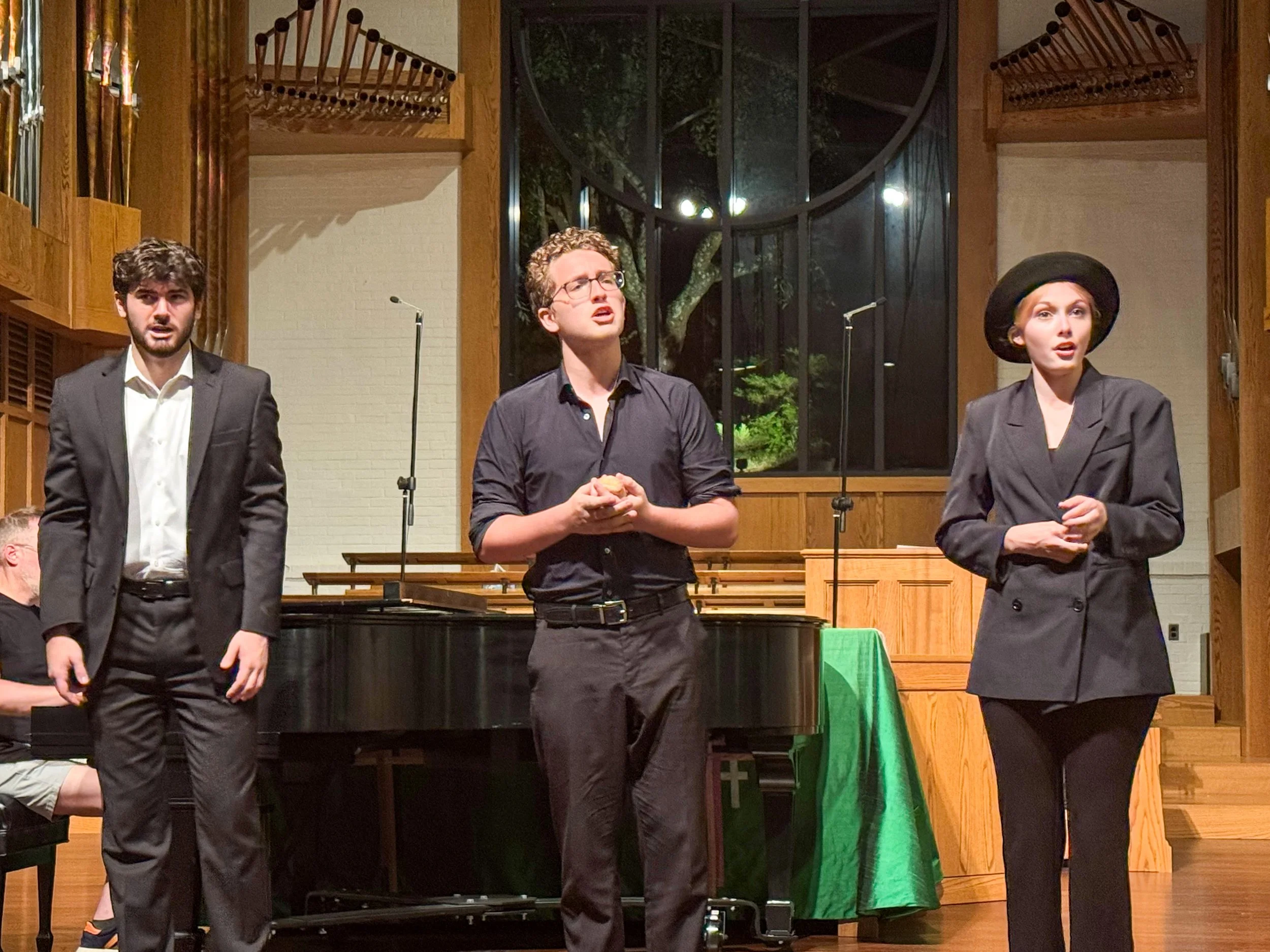 Three performers on stage during a theatrical play, with a pianist in the background, set in a wooden-finished venue with large windows.