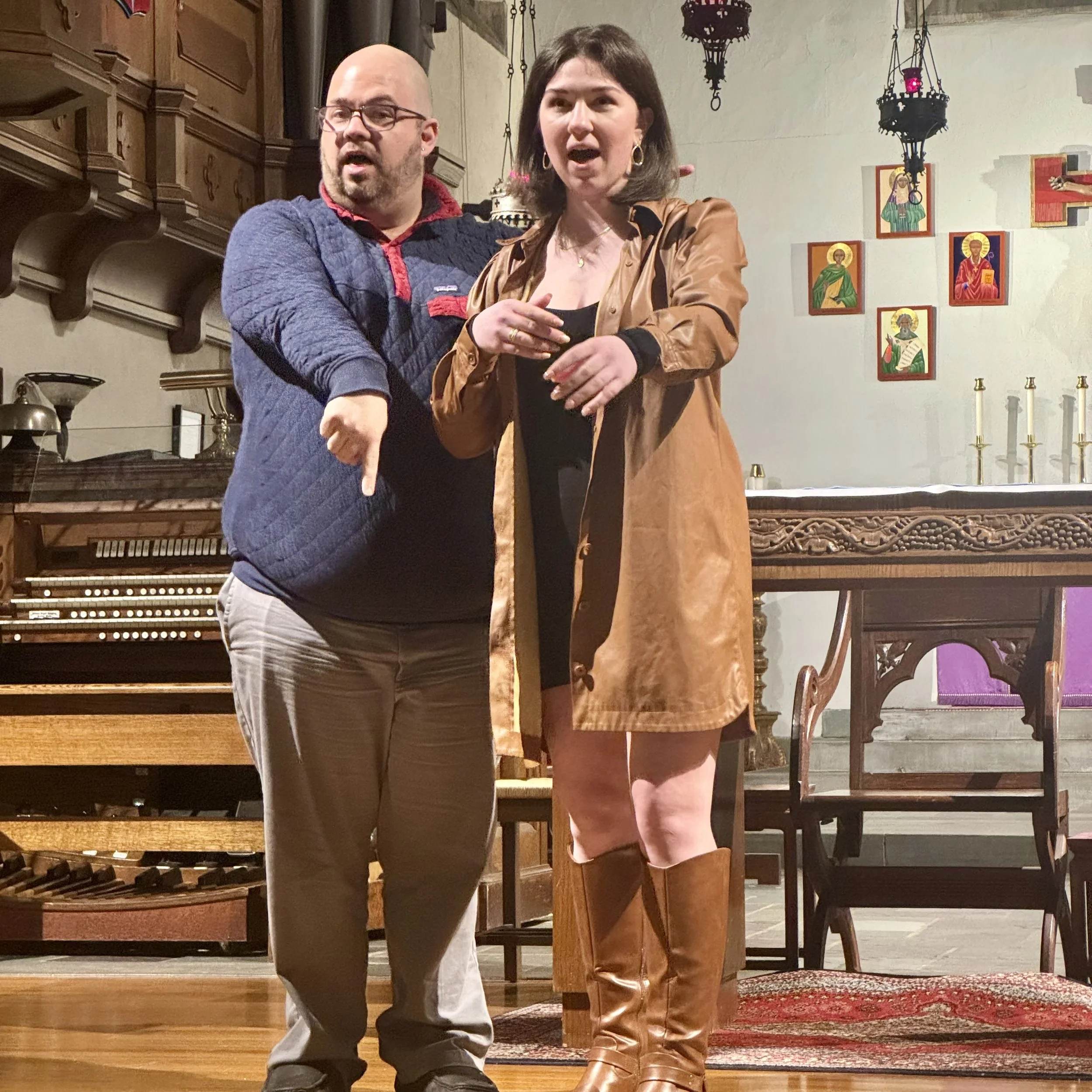 A man and a woman appear to be performing or engaging in a conversation inside a church or chapel, with religious icons and candles in the background.