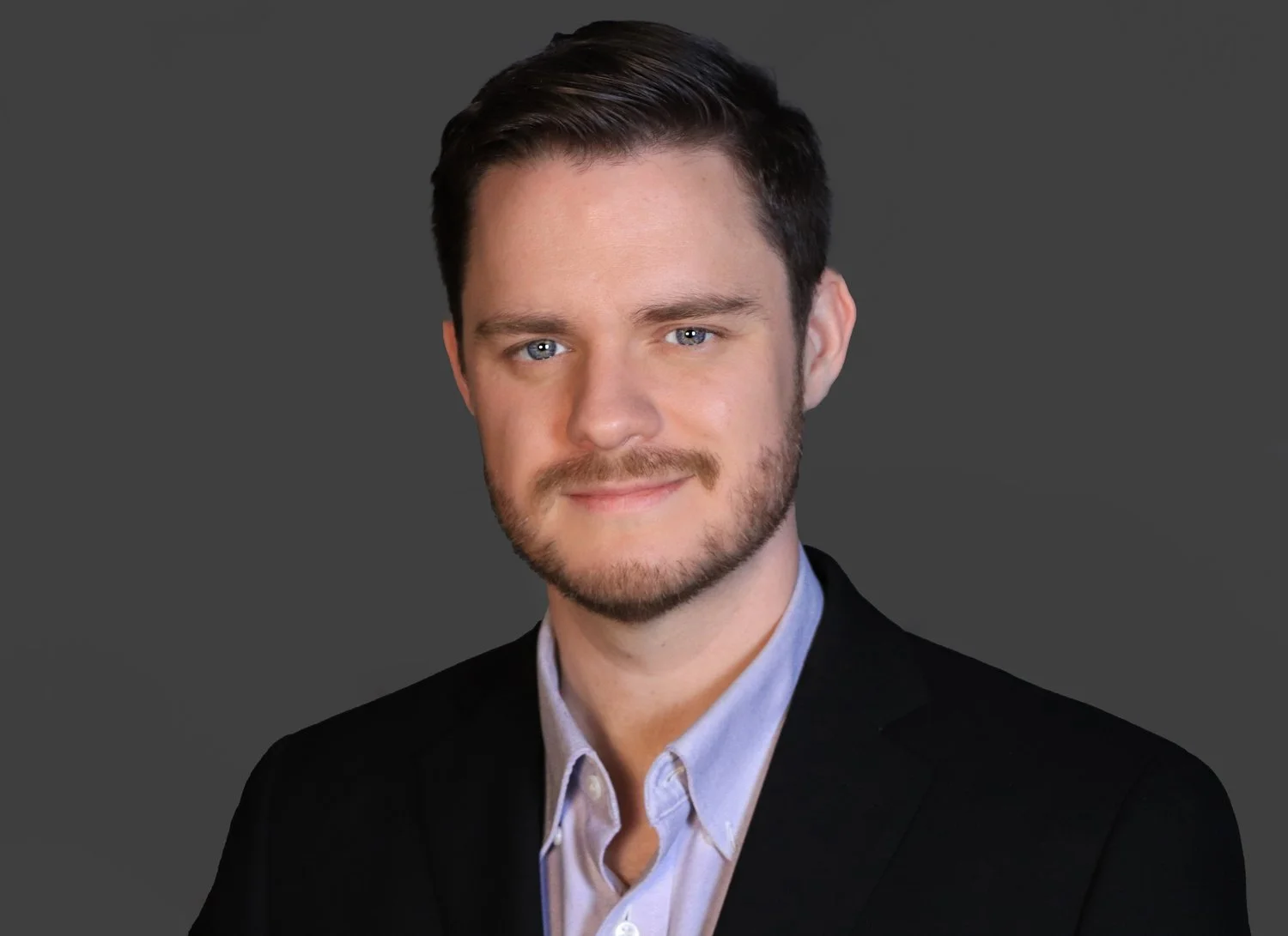 Headshot of a young man with dark hair, blue eyes, beard, wearing a black suit jacket and light blue shirt, against a gray background.