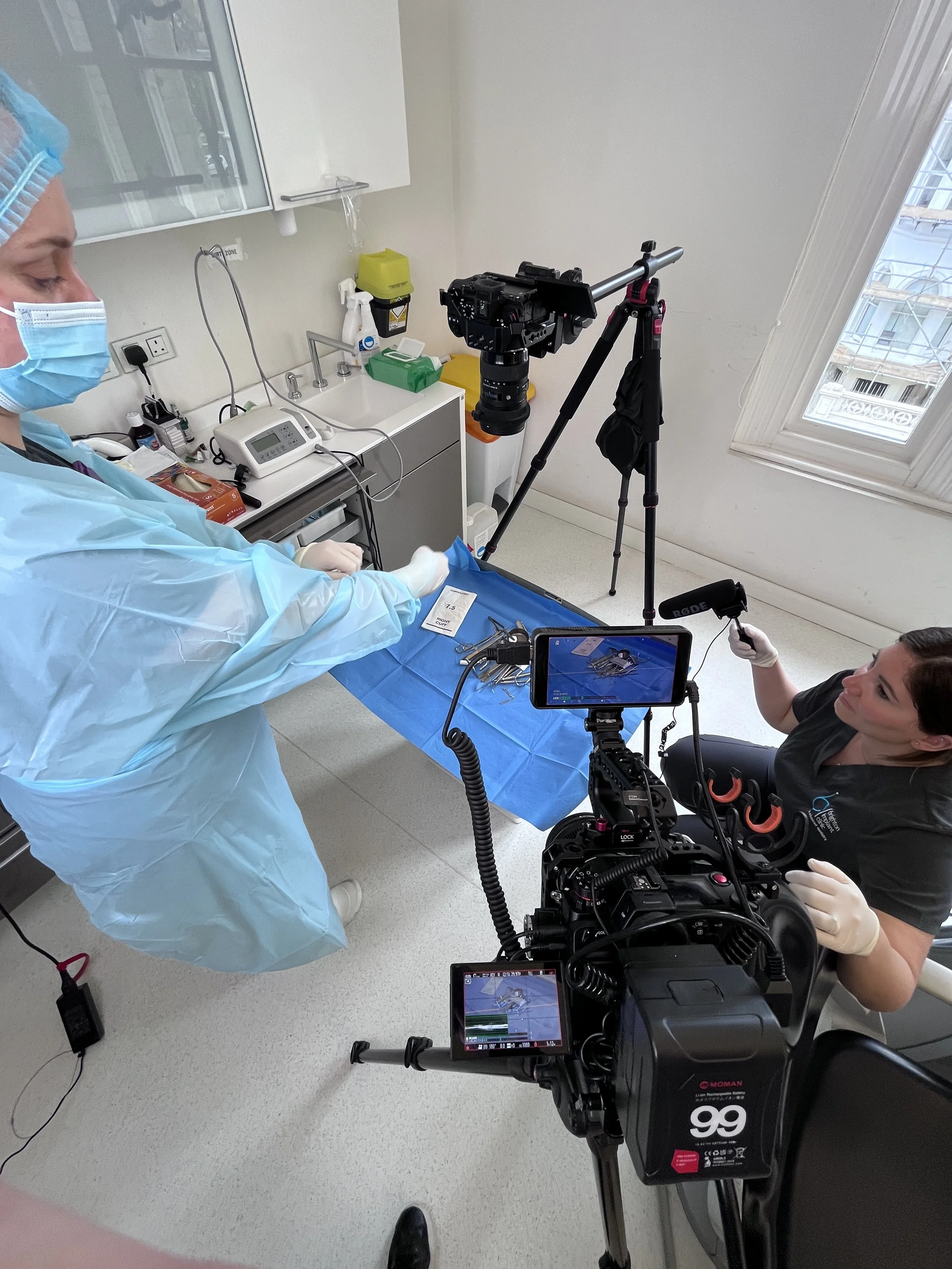 A medical team is filming a procedure in a clinical setting, with a doctor or nurse in full protective gear, including a gown, mask, gloves, and hair covering, working on a patient who is out of frame. Equipment and cameras are set up around the scene, including a large professional camera on a tripod and a smaller camera on a stabilizer, capturing the procedure for documentation or educational purposes.