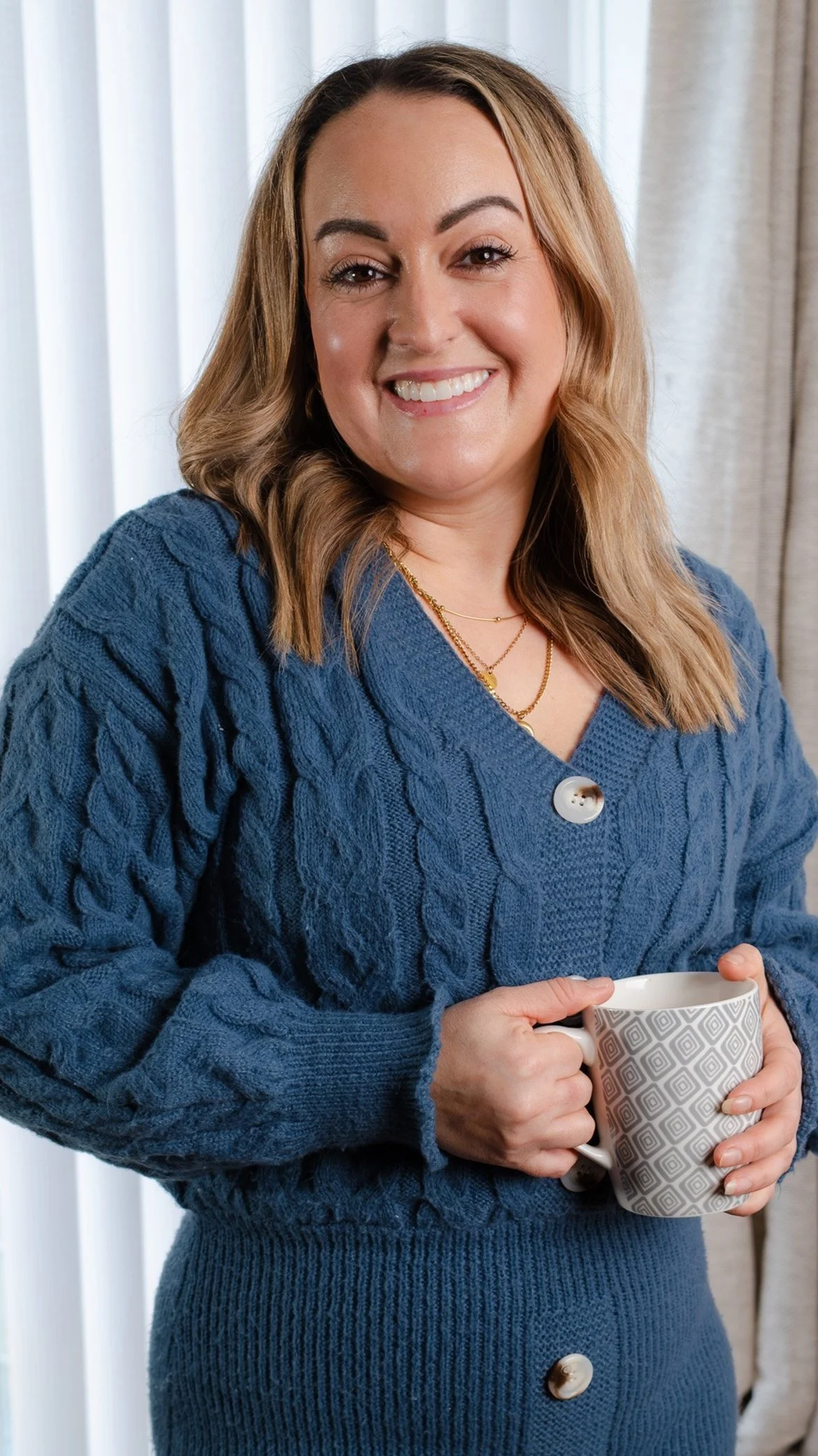 A woman with shoulder-length blonde hair wearing a blue knitted sweater and gold necklaces, smiling and holding a patterned mug with both hands, standing in front of a window with light curtains.
